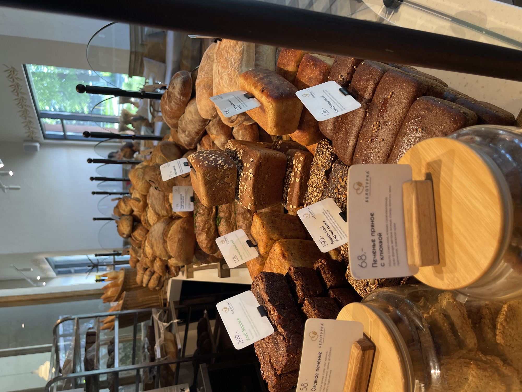 The image shows a display case filled with various types of bread and cookies, likely in a bakery. Each item has a price tag with a description in Russian. The bakery seems to be in Izhevsk, Russia, as the word "БЕЛОТУРКА" (Beloturka) appears on the price tags, which is the name of a local bakery chain. The foreground features jars filled with cookies, while the background shows more bread displayed. A metal shelving unit is seen on the left, possibly for storing additional stock.