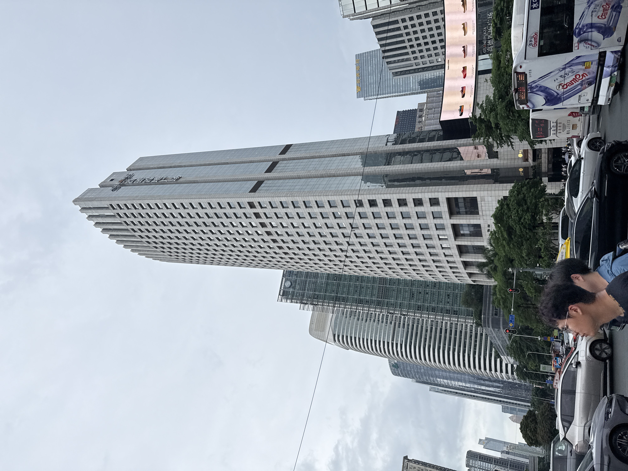 The image captures a street scene in Tianhe District, Guangzhou, China, on an overcast day. A prominent, tall skyscraper with a grid-like facade dominates the center, displaying vertical Chinese characters on its side. To its left, several other modern buildings with curved, layered designs stand in a row. To the right and behind the central skyscraper, other buildings of varying heights and architectural styles are visible. A large digital billboard displaying rows of shoes is mounted on a building. In the foreground, a multi-lane street is filled with traffic, including cars, a white bus with blue and purple branding, and a white minibus. Two young men, one wearing glasses and a black shirt, the other in a blue jacket, are positioned in the lower right corner of the frame, looking towards the right. Traffic lights are visible at an intersection. Lush green trees line the street, interspersed with the urban architecture.
