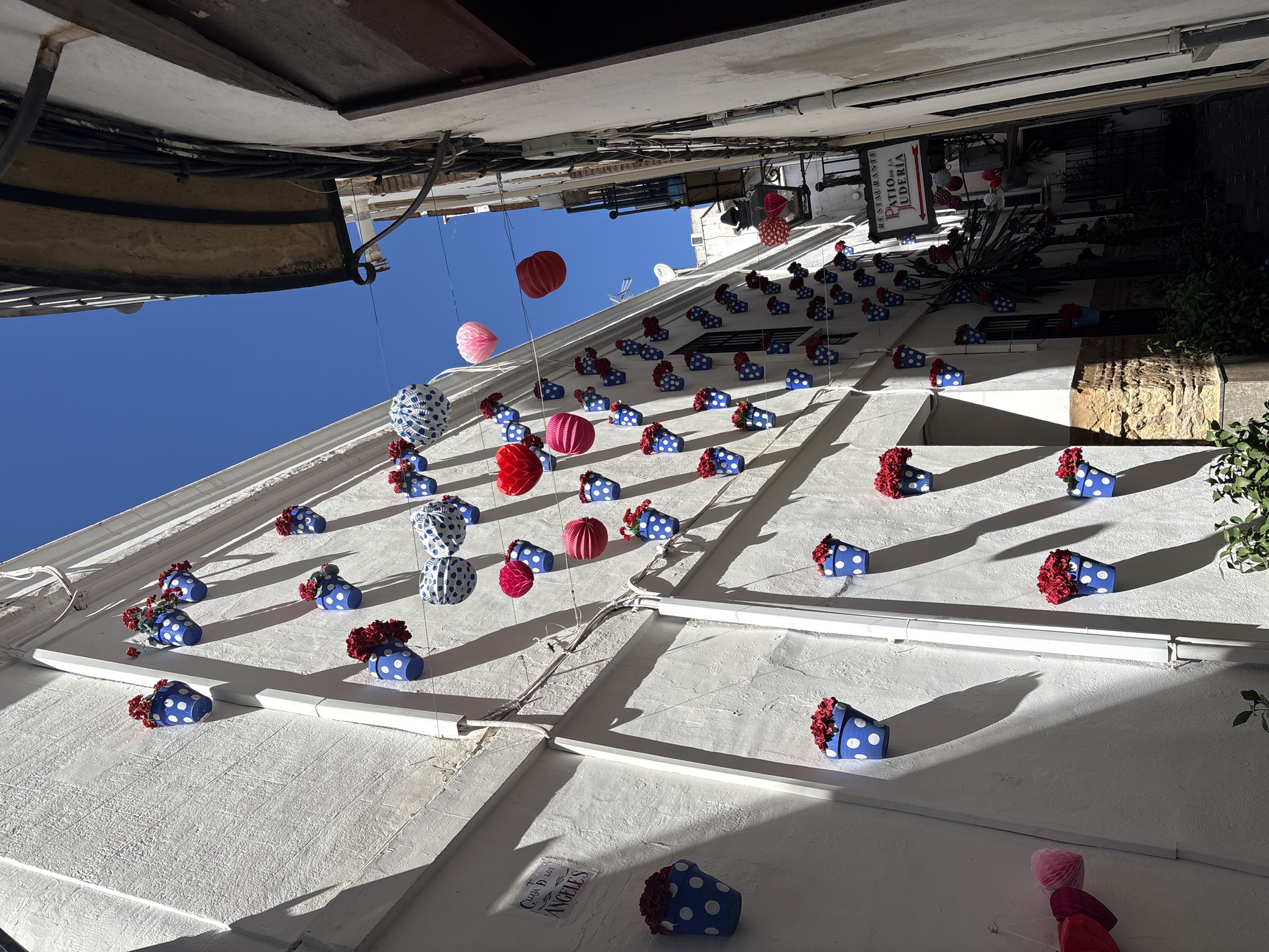 The image captures a street scene in Córdoba, Spain, with a focus on the decorative elements adorning the exterior of a building. The building is painted white, and its façade is decorated with rows of blue polka-dotted vases filled with red flowers. Interspersed among the vases are red and white paper lanterns hanging from above. The street is narrow, and the perspective is from a low angle, looking up at the building and a small patch of blue sky. A restaurant sign is visible towards the top right of the frame, and there are plants hanging near the wall. The scene is lit by sunlight, creating shadows on the building’s surface.