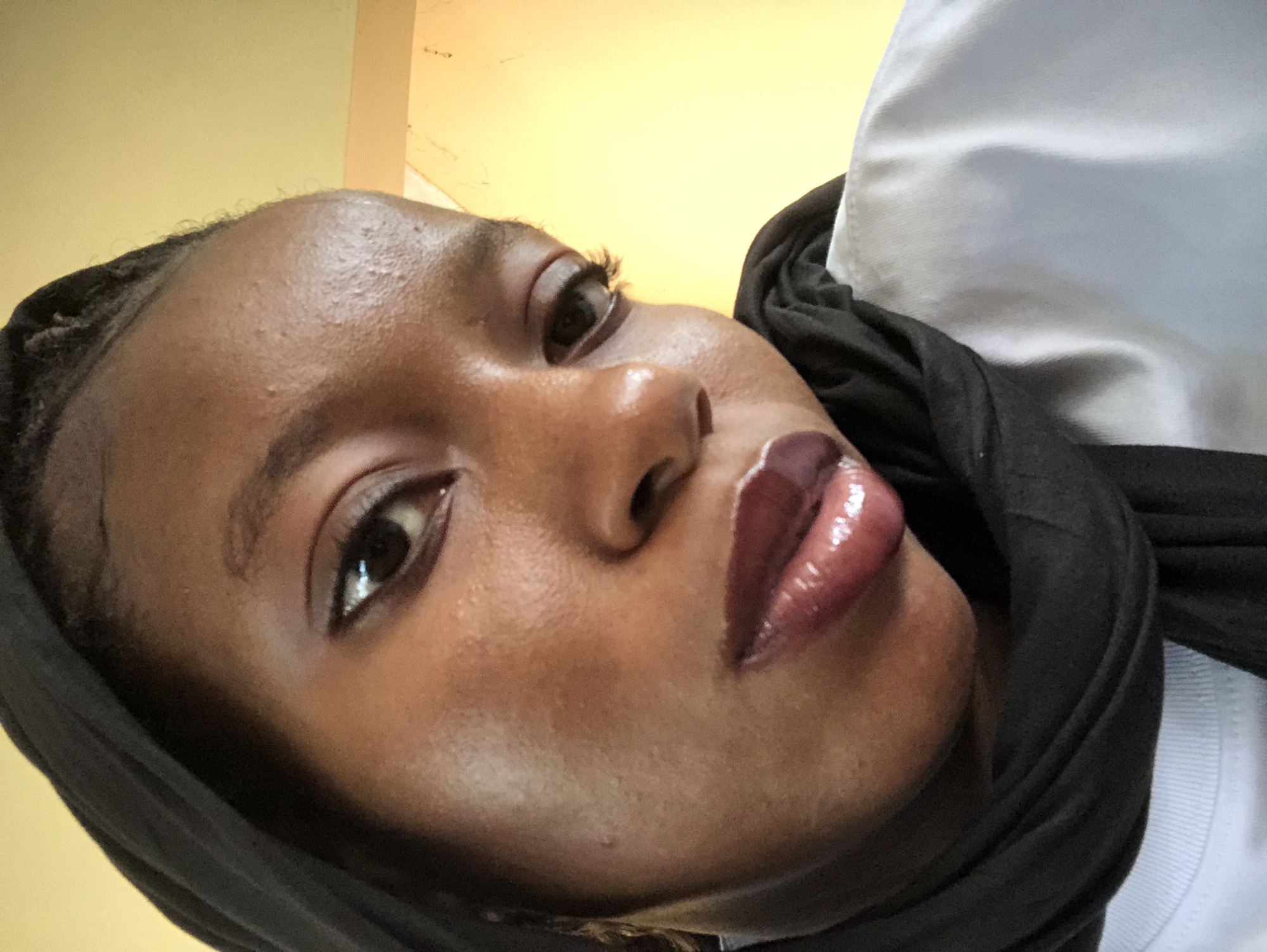 A close-up, low-angle selfie of a dark-skinned person wearing a black headscarf. The person has brown eyes, visible eyelashes, and glossy lips. The background appears to be a light yellow or beige wall. A small portion of a white garment is visible at the top right corner of the frame. There are no obvious indicators to connect this picture with Pindiga, Nigeria.