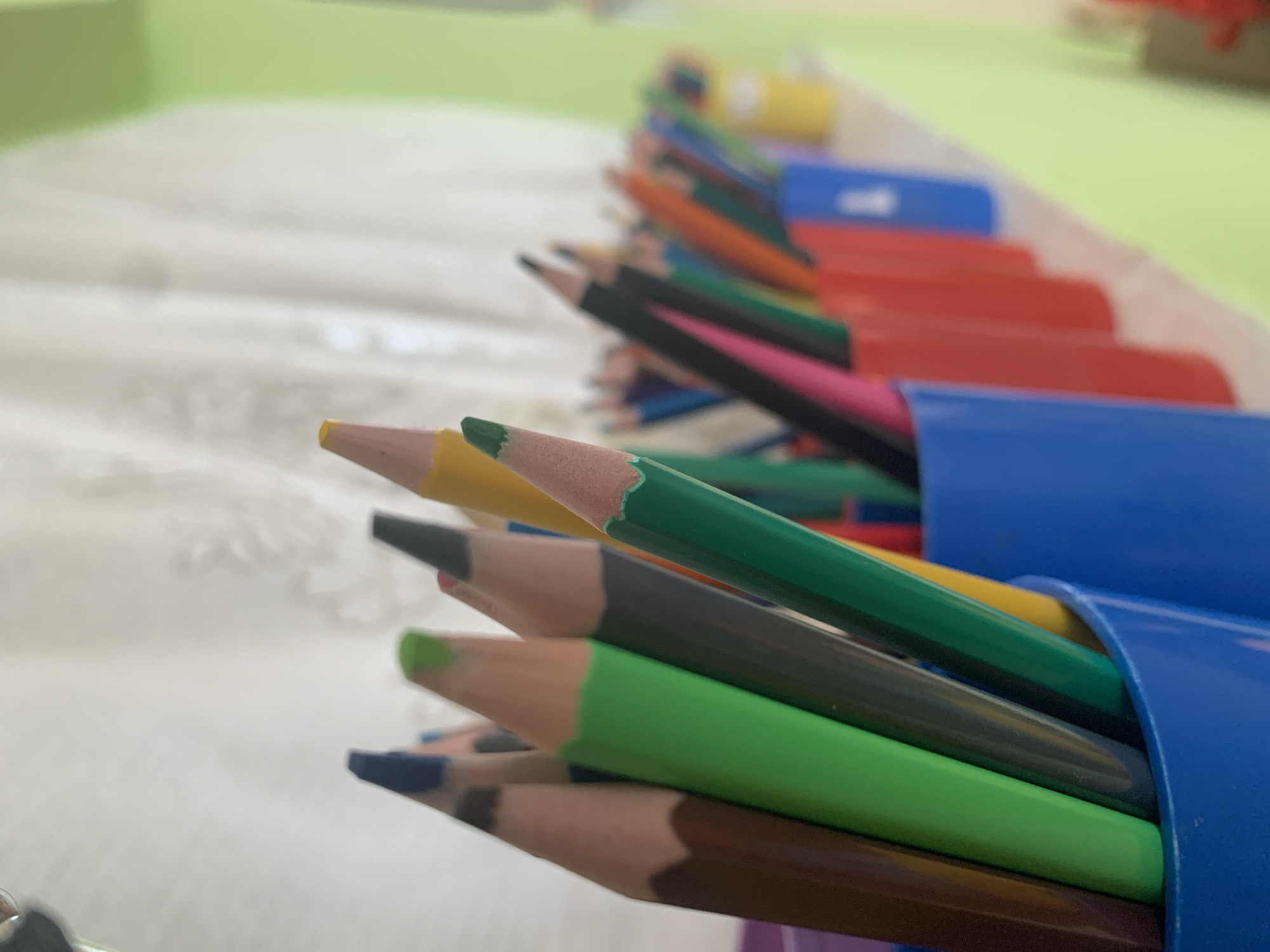 The image shows a close-up view of a collection of colored pencils in blue containers. The pencils are of various colors including green, yellow, brown, black, orange, and red. Some pencils are partially visible in the background. The background is out of focus, but appears to be a white and green surface. The image provides no information to determine a location in Фоки, Russia, or any other city.