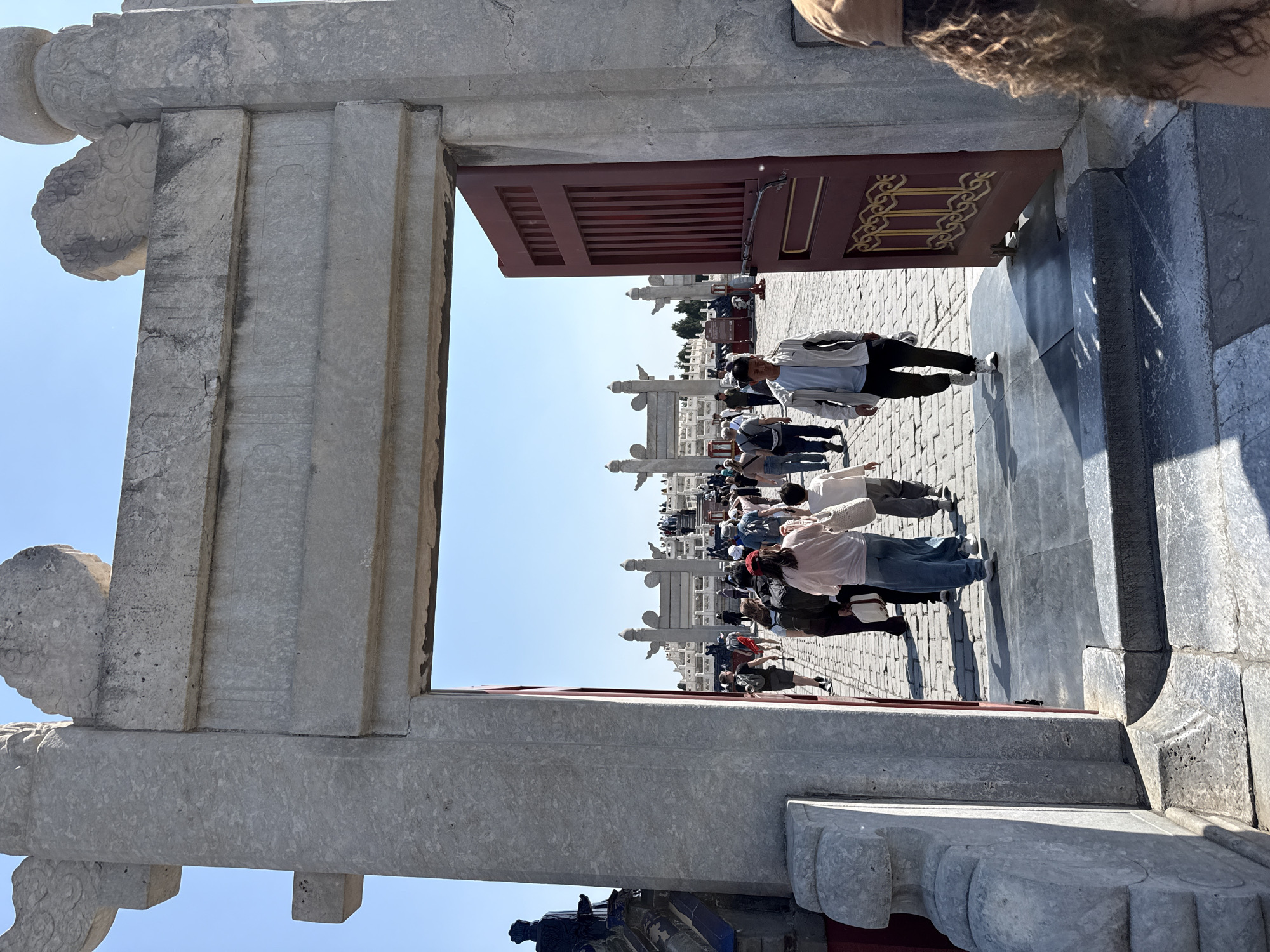 The image captures a scene within the Temple of Heaven in Dongcheng District, Beijing, China. A stone doorway, partially obscured by a red door adorned with golden patterns, frames a view of a paved courtyard bustling with tourists. The doorway is constructed from light-colored stone with detailed carvings. Through the doorway, multiple people are visible, walking in various directions. Stone balustrades line the courtyard, adding to the architectural detail. The sky above is clear and blue.