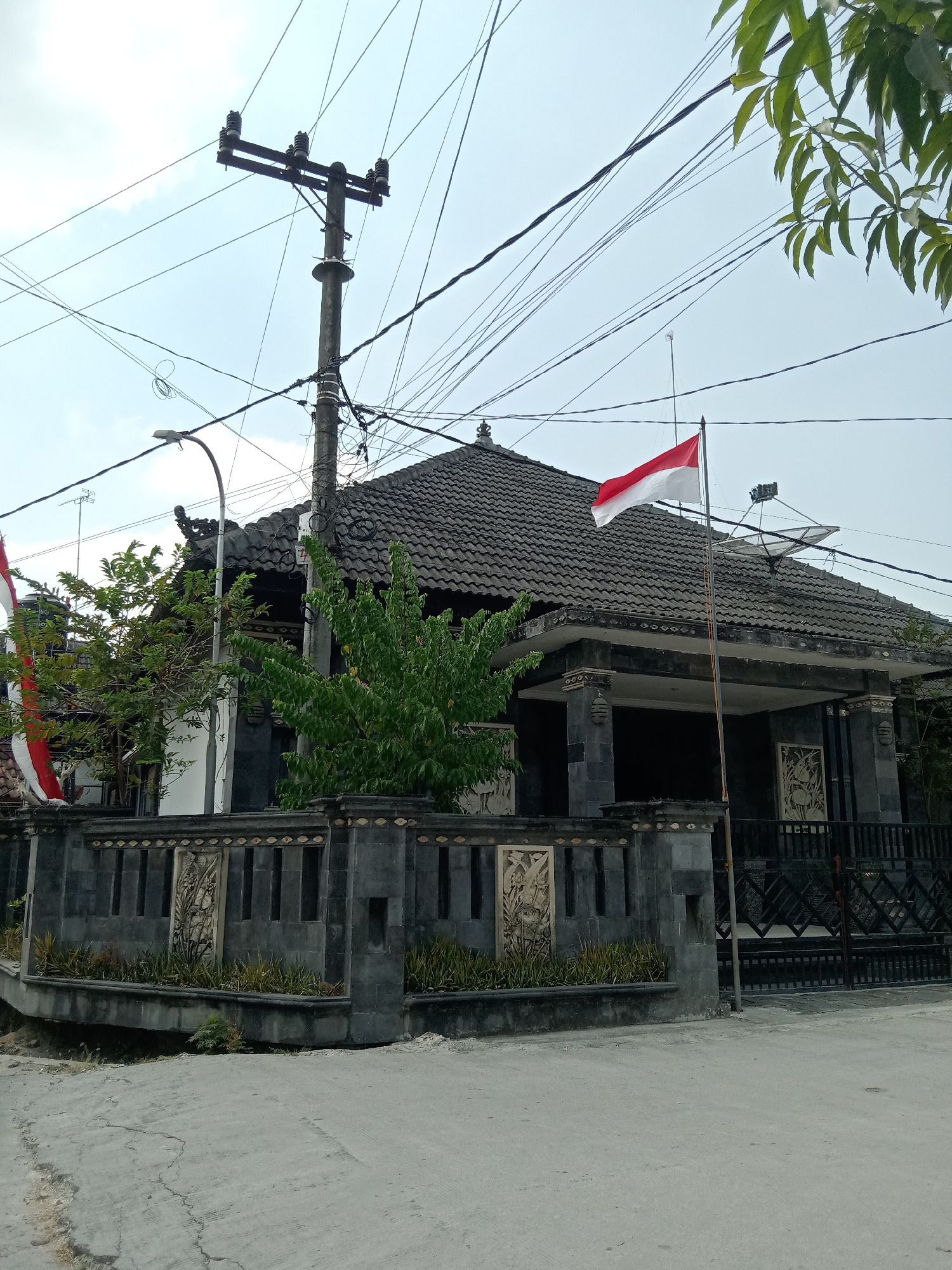 In the city of Jumput, Indonesia, a low-angle shot captures a traditional Balinese house with a dark tiled roof. The house features ornate stone carvings on its exterior walls and a covered porch supported by dark columns. In front of the house, a gray stone fence with decorative inlays lines the property. An Indonesian flag, red and white, flies from a flagpole in front of the house. Overhead, a complex network of electrical wires and a utility pole crisscross the sky, with a street lamp also visible. Lush green foliage from trees partially obscures the house and fence. The foreground shows a cracked concrete road.