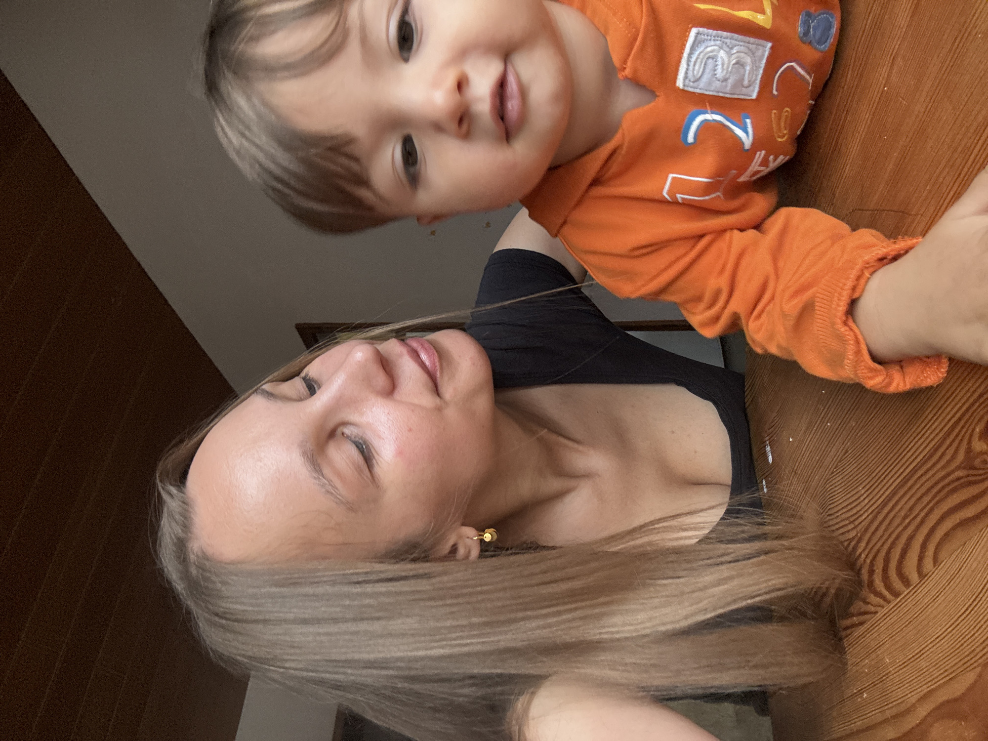 In a close-up indoor shot, a woman with long blonde hair and a black top looks to her left, her gaze directed slightly downwards. She has a subtle smile on her face and is wearing a small gold earring. Beside her, a toddler with brown hair, wearing an orange hooded sweatshirt with white and blue numbers and designs, looks towards the camera. They are both seated at a wooden table with a distinct wood grain pattern. The background is blurred, showing a dark brown wooden ceiling and a light-colored wall. The scene appears to be a domestic setting, likely a home environment in Überherrn, Germany.
