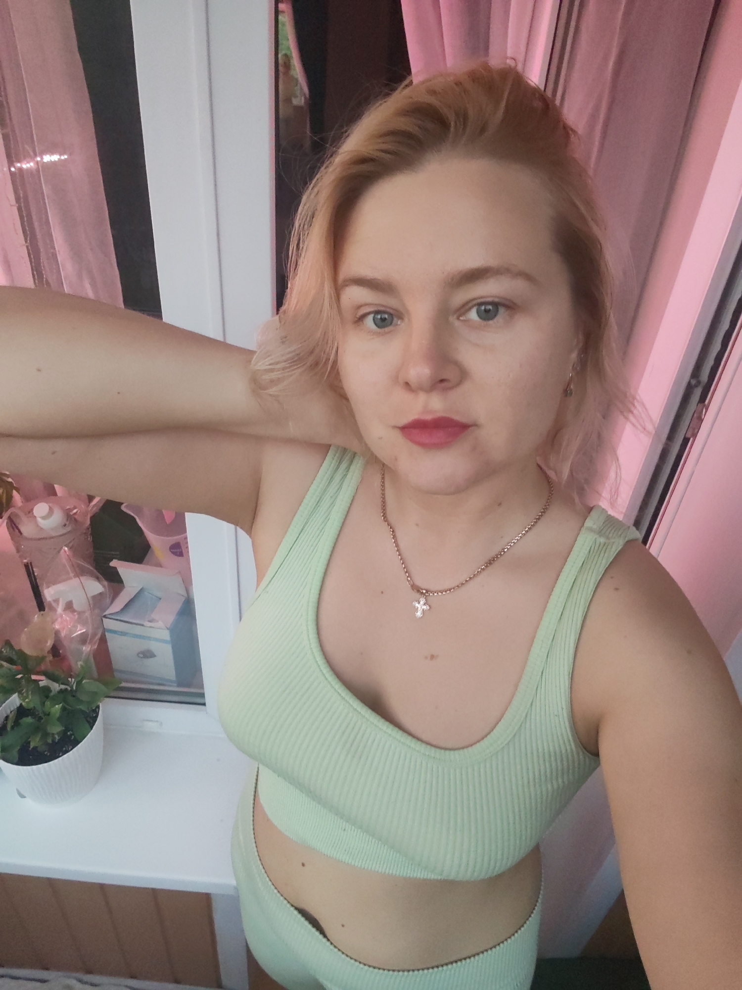 A young Caucasian woman with blonde hair and blue eyes is taking a selfie indoors. She is wearing a light green ribbed crop top and matching bottoms. She has her right arm raised and resting behind her head, with her left hand near her neck. A silver chain with a cross pendant is visible around her neck. Behind her, a window with sheer pink curtains is visible, along with a small potted plant on the windowsill. The overall setting appears to be a domestic environment. The image does not provide any specific information about the city of Izhevsk, Russia.