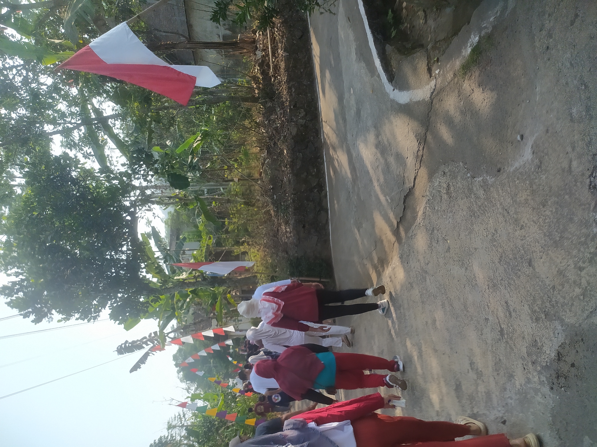 In the village of Lopait, Indonesia, a group of people walks along a concrete road lined with trees and flags. The red and white Indonesian flag is prominently displayed on a pole to the right, and a string of smaller red, white, and multi-colored flags stretches across the road in the background. The people, dressed in casual attire, appear to be participating in a community event or procession. The sun casts long shadows on the road, indicating it is likely morning or late afternoon. The surrounding environment is lush and green, with dense foliage and what appears to be a small building partially visible behind the trees.