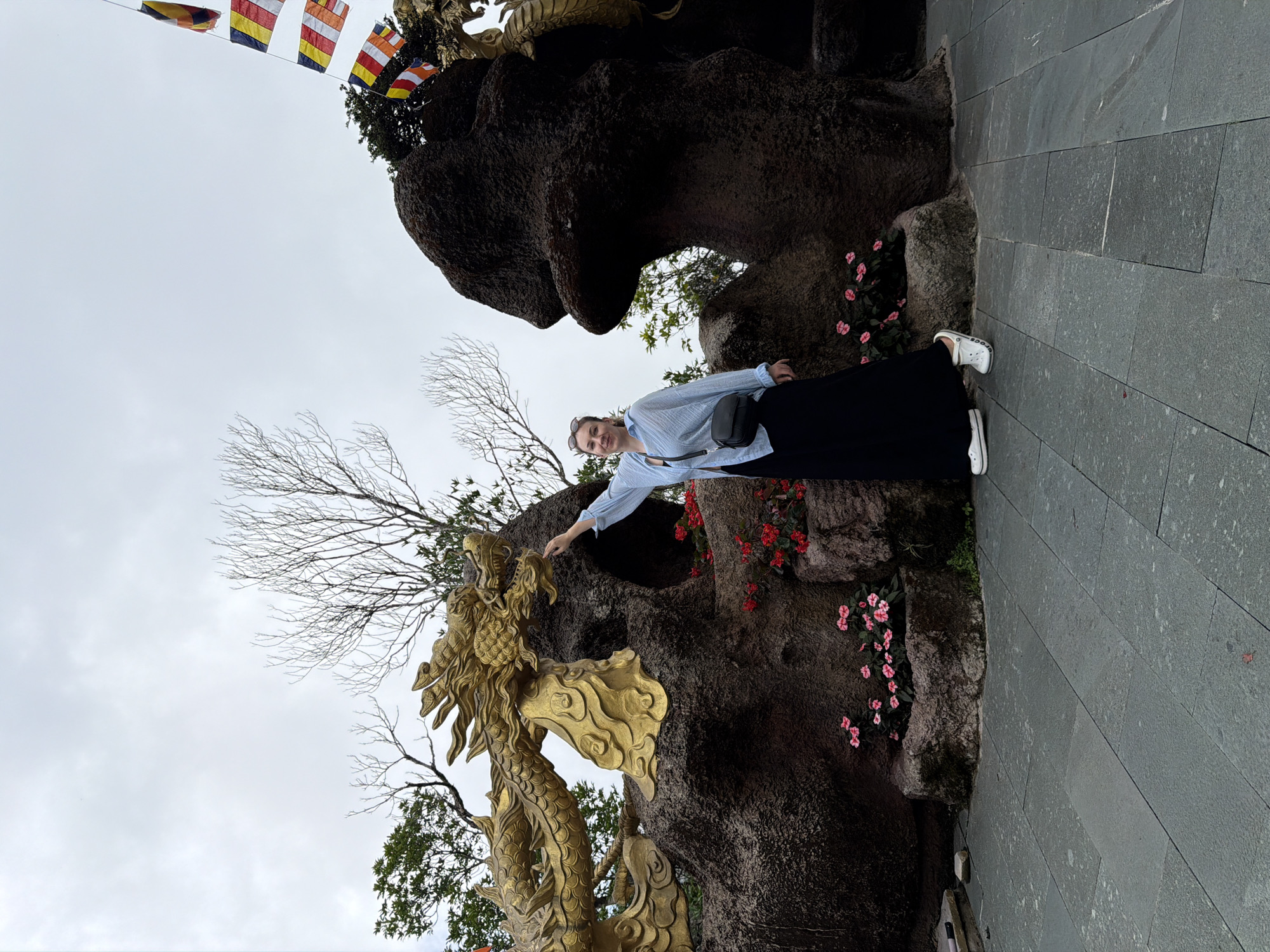 A woman stands in front of a large, dark, rock-like structure, with a golden dragon sculpture to her left. The dragon is coiled and detailed, with scales and a fierce expression. The woman is wearing a light blue, long-sleeved shirt and a long black skirt. She has a black cross-body bag and white sneakers. She is smiling and has her right hand extended towards the dragon. In the background, there are some trees and colorful Buddhist flags hanging from poles. Red and pink flowers are planted around the base of the rock structure. The ground is paved with large, grey stone tiles. The sky is overcast. This scene is located at the Linh Ung Pagoda on Son Tra Peninsula in Da Nang, Vietnam.
