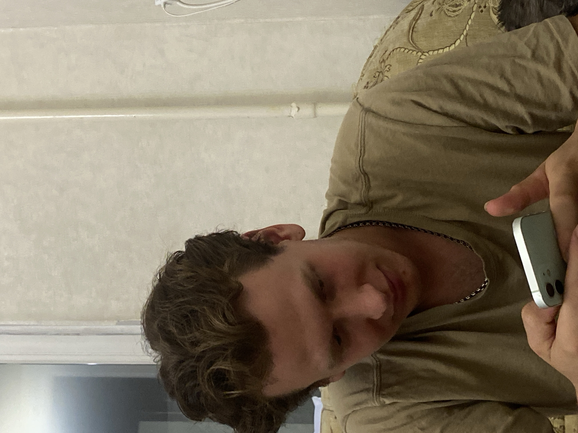 The image shows a young man with curly brown hair, wearing a beige t-shirt and a silver chain, looking down at a smartphone he is holding in his hands. He appears to be indoors, with a textured wall in the background and a glimpse of a patterned sofa on the right. The lighting suggests an indoor setting, possibly a residential interior. There are no clear indicators of the specific location within Bishkek City, Kyrgyzstan.