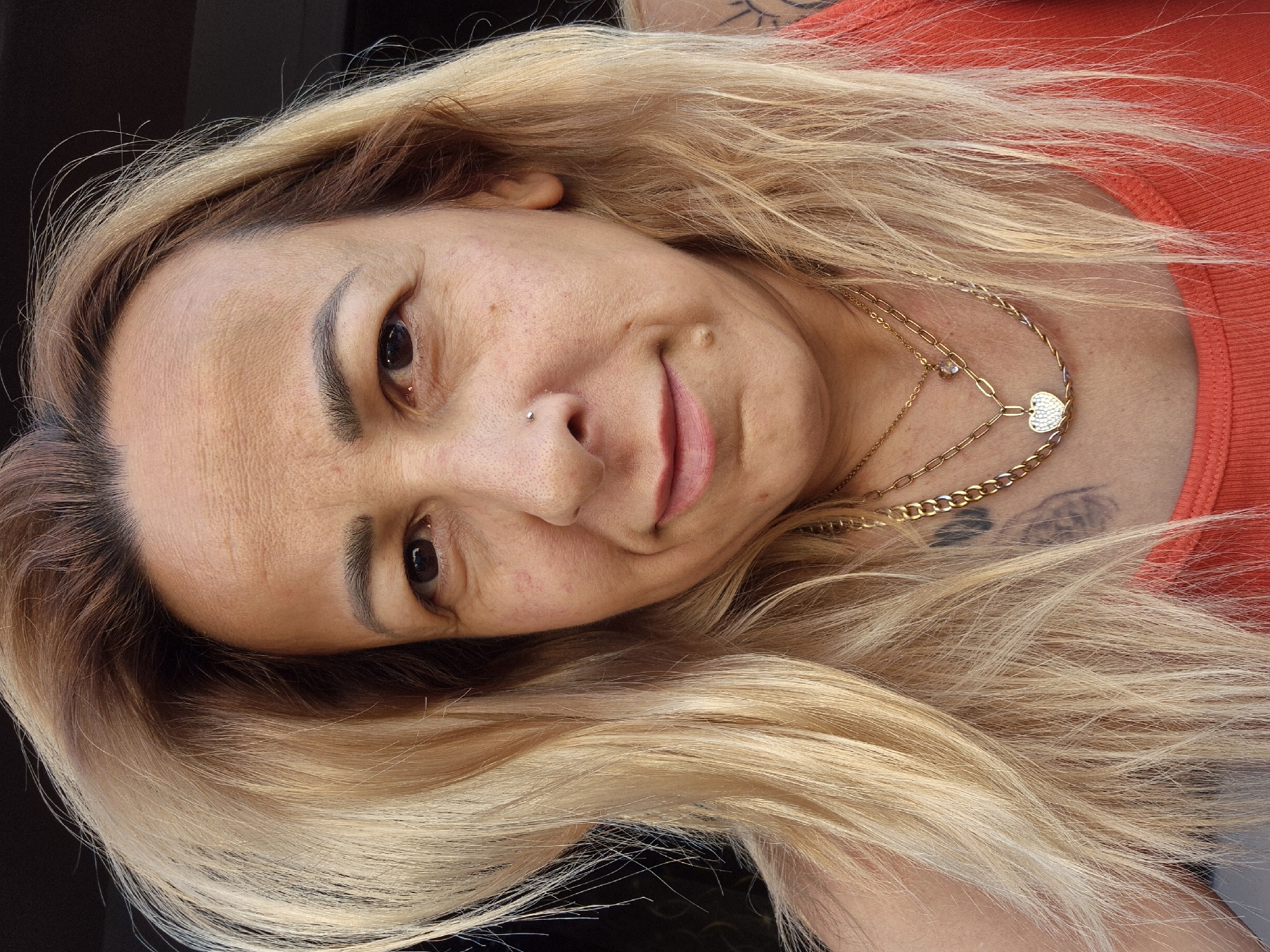 A close-up, over-the-shoulder selfie of a woman with blonde hair, who is looking directly at the camera. She has fair skin, light brown eyes, and a small nose piercing. She is wearing an orange tank top and multiple gold necklaces, one of which has a heart-shaped pendant. Tattoos are visible on her left shoulder and chest. The background is dark and out of focus.