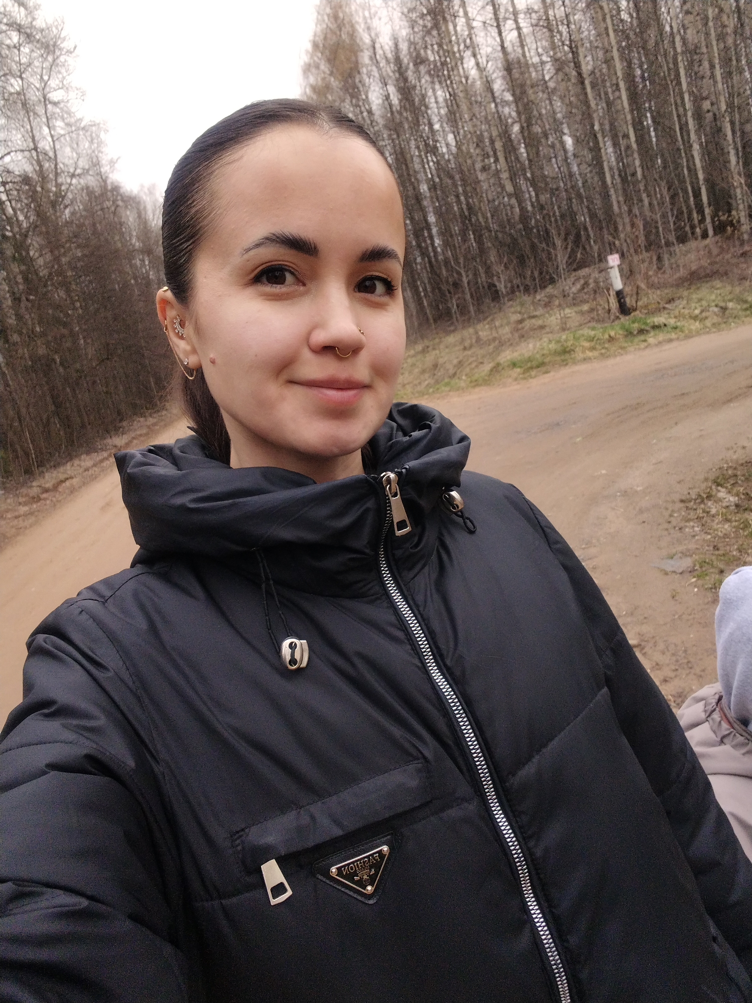 A young adult female is featured in an outdoor, close-up selfie, smiling gently at the camera. Her dark hair is pulled back, revealing a nose ring and multiple piercings on her left ear, including a decorative earring with small stones and a chain connecting two studs. She is wearing a black, puffy winter jacket with a prominent silver zipper and a silver toggle on the drawstring near the neck. A black triangular patch on the jacket displays the text "FASHION" (appearing reversed) and "MILANO," styled similarly to a well-known luxury brand logo.
The setting is a rural or forested area, characterized by a dirt or gravel road. A dense forest of bare, deciduous trees lines the background, suggesting either late autumn or early spring. The sky is overcast, indicating a cool, cloudy daytime. A white and red striped pole or marker is visible amidst the trees. Part of a gray hoodie is visible on the right side of the frame, hinting at the presence of another person. The scene aligns with a natural outdoor environment, possibly near Noviy, Russia.