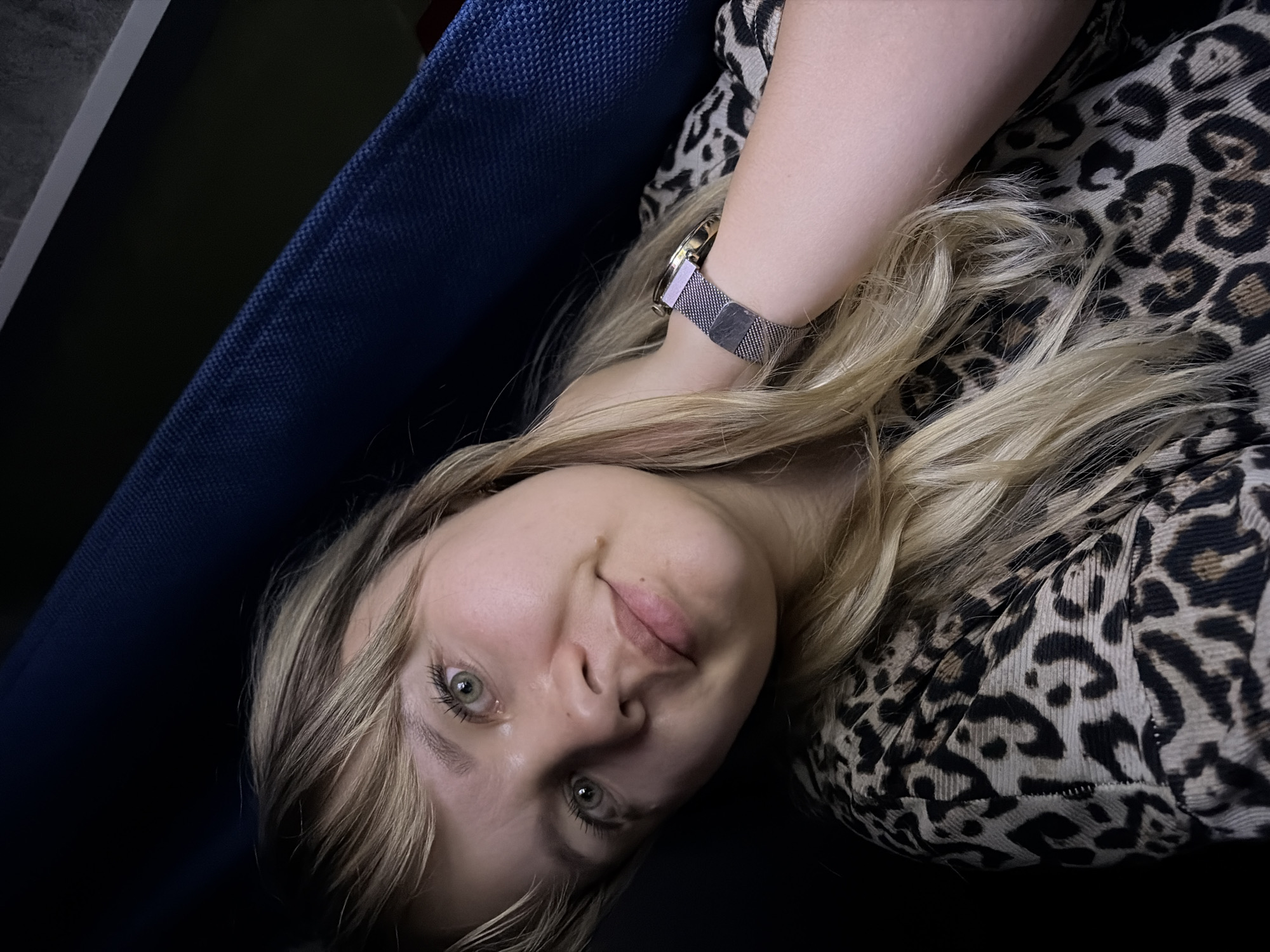 A woman with light blonde hair and green eyes is reclining on a blue fabric surface. She is wearing a leopard-print top and a metallic wrist watch. The location is unknown and there are no indications that it is Chaikovsky, Russia.