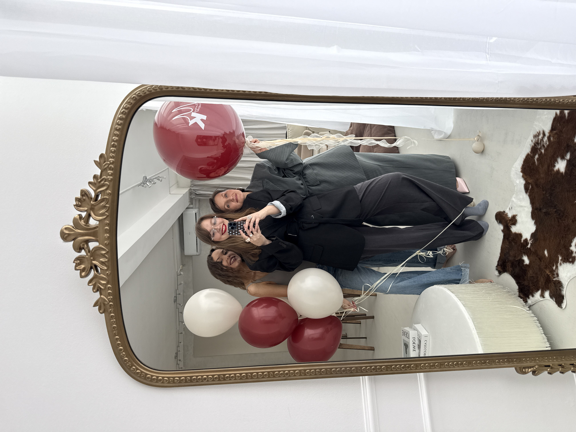 The image shows a reflection in an ornate, gold-framed mirror. Three women are taking a selfie. Two red balloons and two white balloons are visible in the mirror. Behind the women, the reflection shows furniture including a striped chair or couch, a white tufted pouf with books on it, and a cowhide rug. The location, Izhevsk, Russia, cannot be verified.