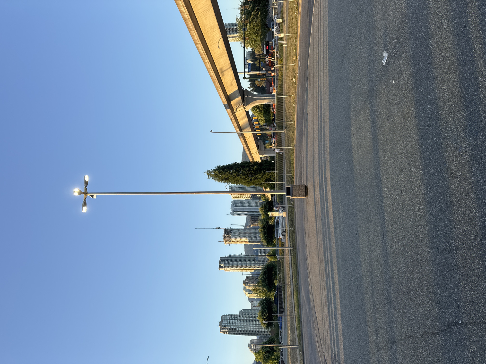 The image depicts an outdoor urban scene in Coquitlam, Canada, likely during a sunny late afternoon given the shadows. In the foreground, a vast asphalt parking lot stretches towards the viewer, with a prominent streetlamp with four heads positioned in the center. A dark trash receptacle stands at the base of the streetlamp. The middle ground is characterized by a chain-link fence separating the parking lot from a grassy area and a road. Beyond the fence, a concrete elevated structure, likely a SkyTrain line, dominates the upper right portion of the frame, casting long shadows. A line of trees partially obscures a distant cityscape dominated by several tall, modern buildings and construction cranes. The sky is clear and blue. The scene appears quiet with no people or vehicles actively engaged in the immediate foreground or mid-ground.