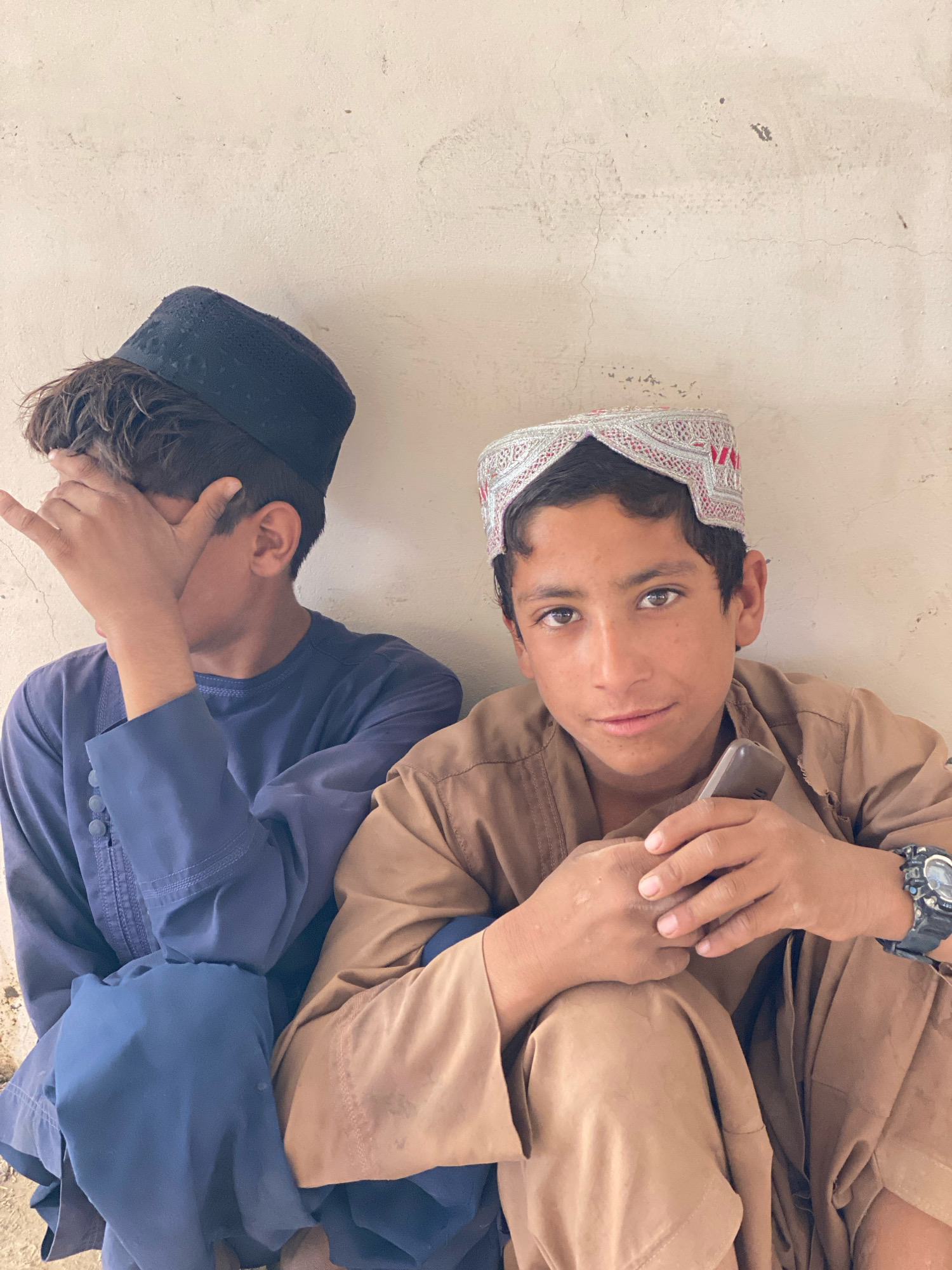 Two young males are sitting against a textured, light-colored wall. The male on the left, partially obscuring his face with his hand, wears a dark skullcap and a deep blue tunic with decorative stitching and buttons. The male on the right, looking towards the camera, wears a white, intricately embroidered cap with red accents and a light brown tunic. He is holding a mobile phone in his right hand and has a wristwatch on his left wrist. The environment appears to be outdoors with an unadorned wall providing the backdrop. The scene suggests a candid moment, possibly in the city of Pasab, Afghanistan, given the attire and cultural context.