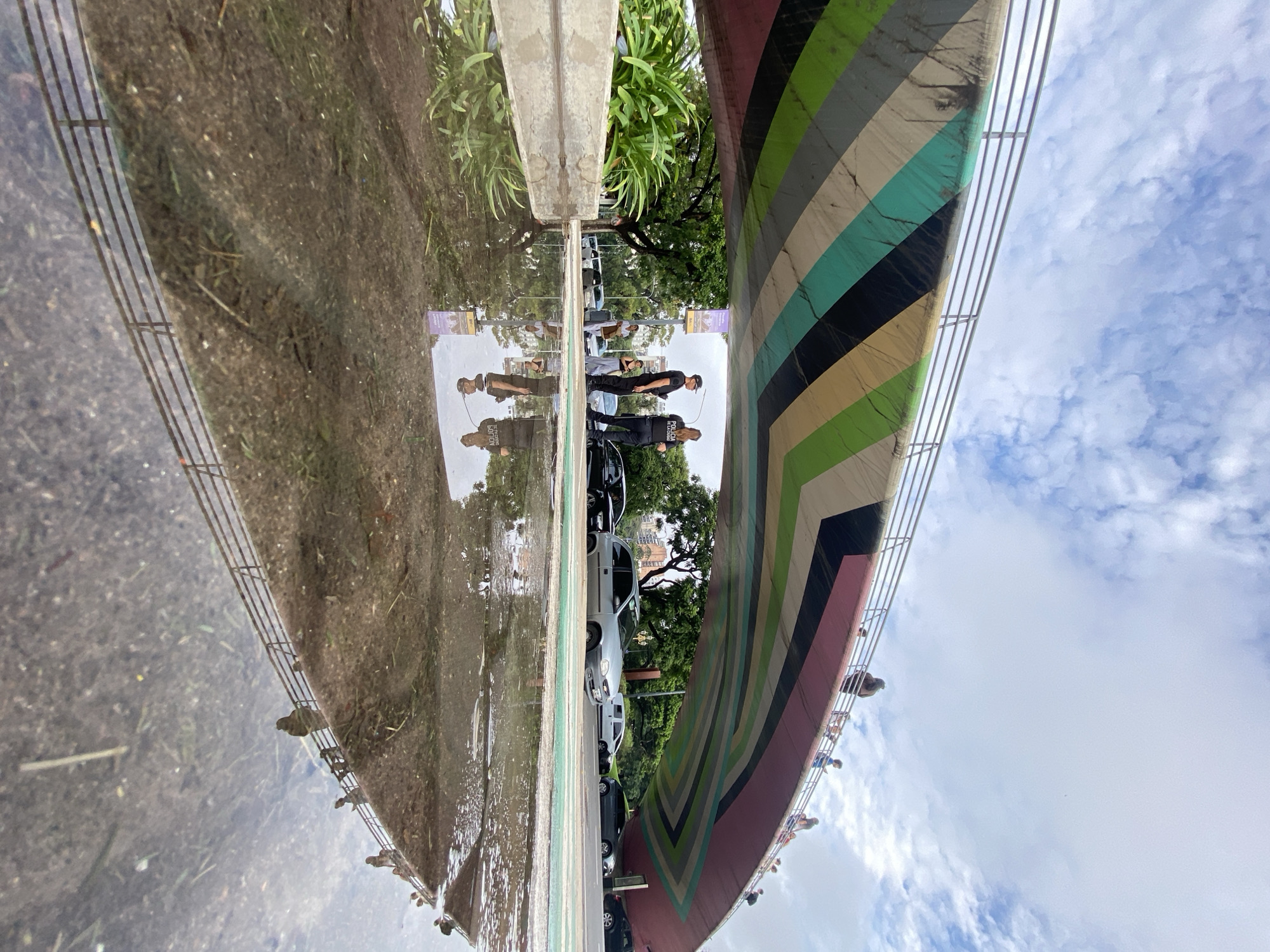 This is a photograph of the Vuelta de Rocha bridge in Buenos Aires, Argentina, reflected in a puddle. The bridge is painted with colorful stripes. There are people walking on and near the bridge. Cars are parked alongside the bridge. The sky is visible with clouds. A concrete structure and some vegetation are also reflected in the puddle.