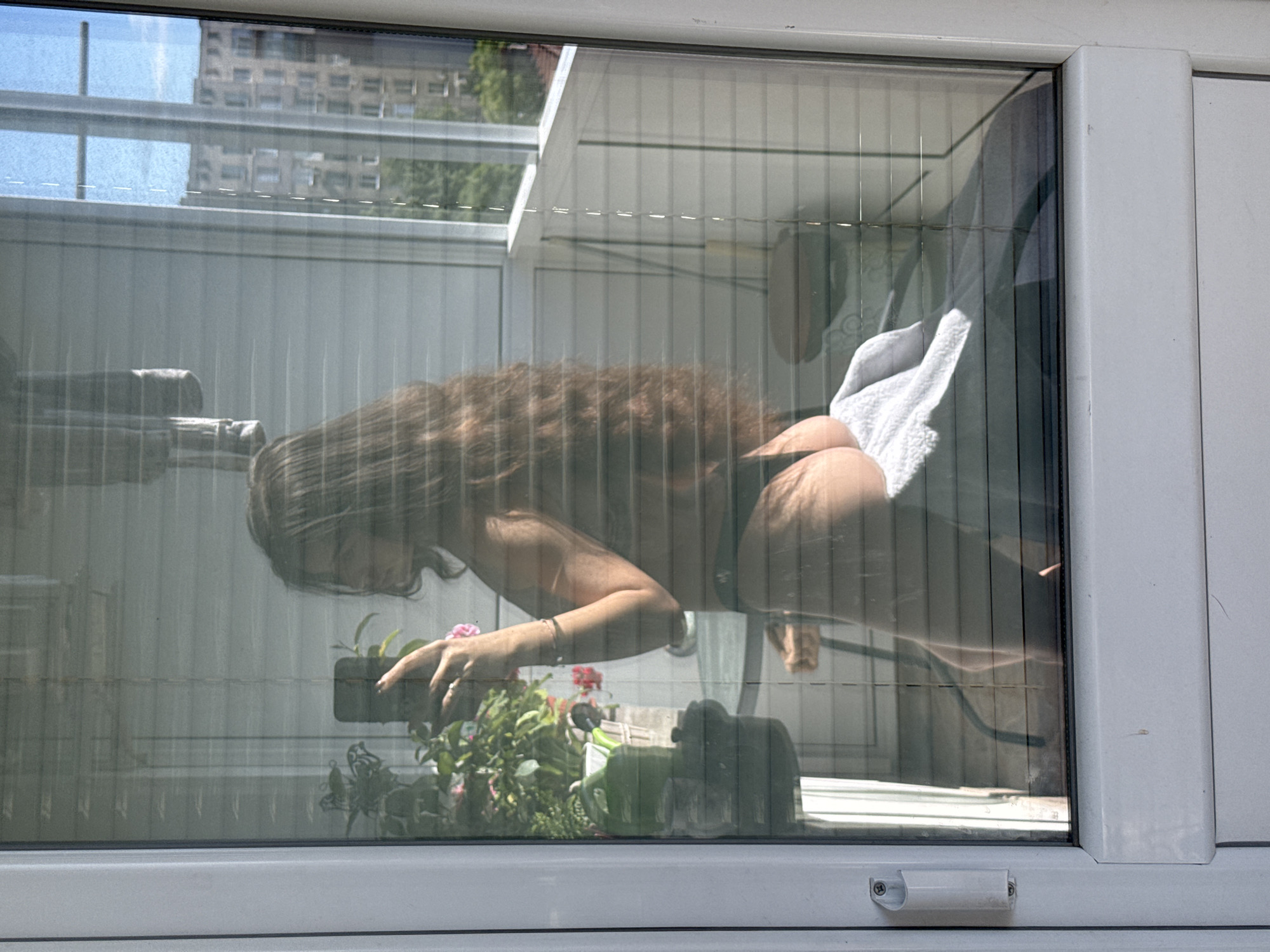 A woman with long, brown hair is seen through a window. She is bent over, her left hand touching a potted plant. A white towel is partially visible behind her. The image appears to be taken from outside, looking into an interior space. The background includes a building and what seems to be a balcony. The location in Plovdiv, Bulgaria is not discernible from the image.