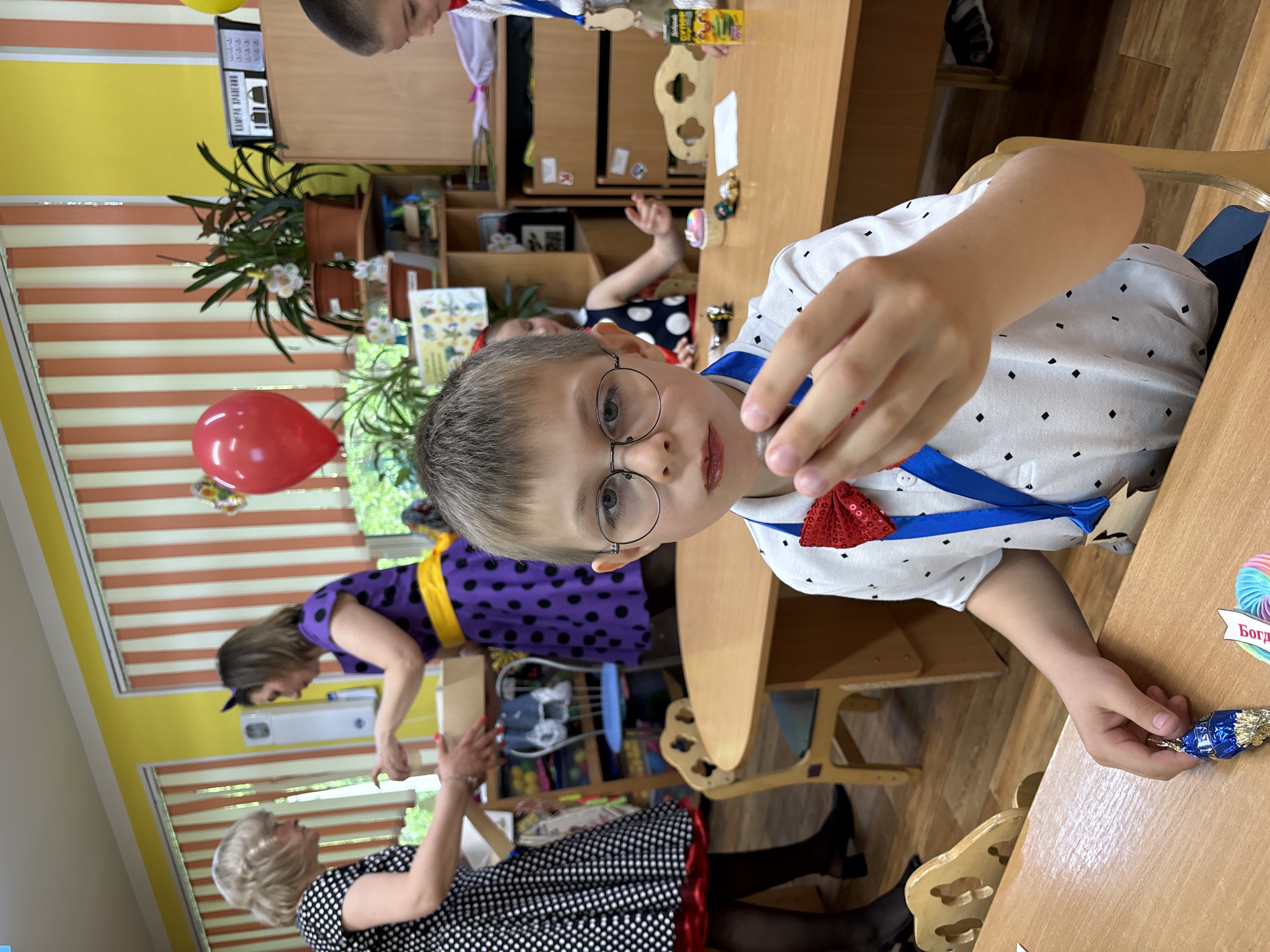 A young boy with glasses sits at a wooden desk, dressed in a white shirt with black diamond patterns and a red bow tie, holding a wrapped candy in his hand. He has a blue ribbon around his neck with a star-shaped decoration attached. A red balloon is visible in the background. Two women are standing near a shelf filled with various items. One woman is wearing a black and white polka dot dress with a red trim. The other woman is wearing a purple dress with yellow accents. The location appears to be indoors, possibly in a school or daycare setting.