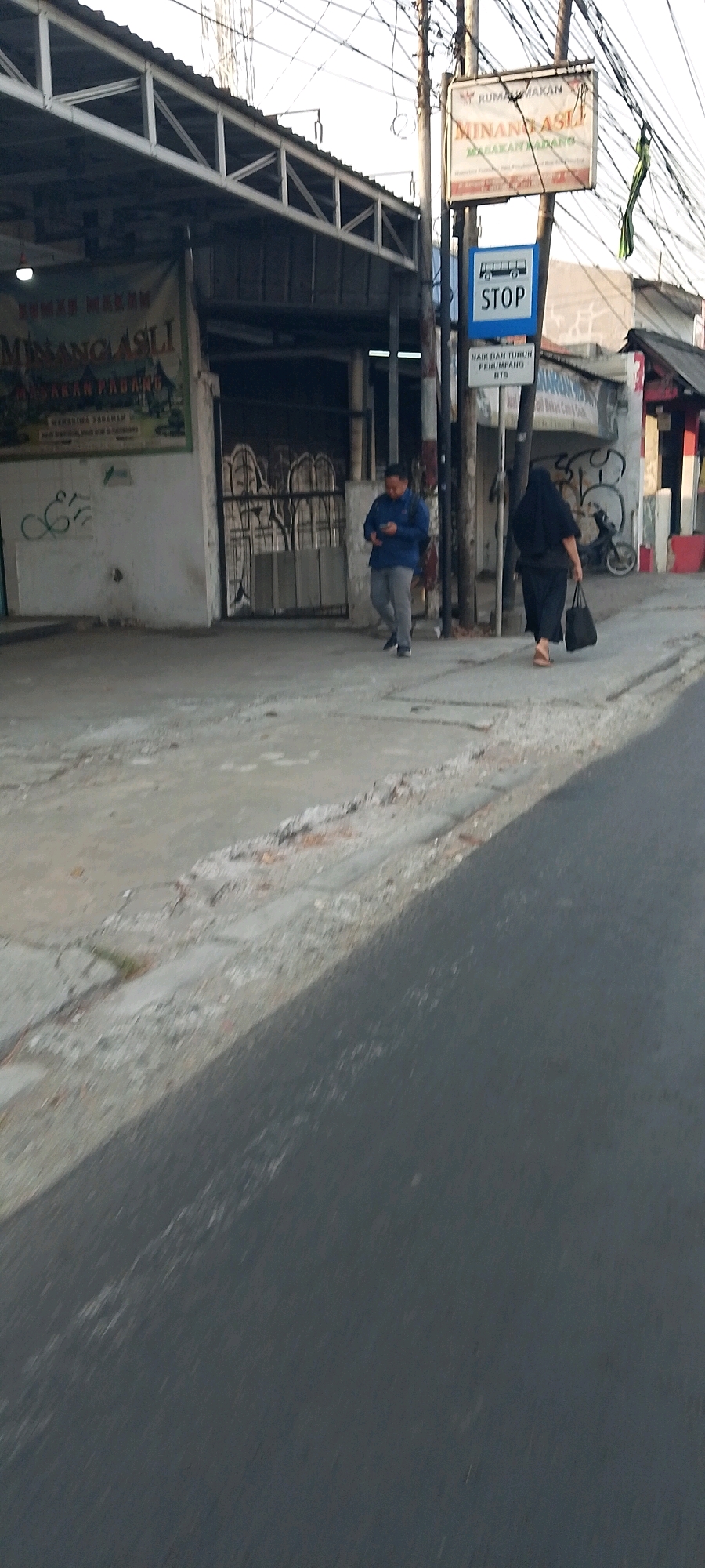 The image depicts a street scene in front of a building with a covered walkway, likely in the city of Bekasi, Indonesia, as indicated by the signage. The building appears to be a restaurant, with a banner advertising "RUMAH MAKAN MINANGKABAU ASLI" (Authentic Minangkabau Home Cooking). A blue sign with a bus icon and the word "STOP" suggests a bus stop. Another sign below it reads "NAIK DAN TURUN PENUMPANG BTS" (Pick up and drop off passengers BTS).
Two individuals are visible. On the left, a man in a blue jacket and grey pants walks while looking at his phone. On the right, a woman in a black headscarf and black clothing walks away from the camera, carrying a black bag. A motorcycle is parked in the background. The ground is a concrete pavement with visible cracks and some debris. The overall environment appears to be a commercial area with overhead wires and a somewhat weathered appearance of the buildings. The lighting suggests it is daytime, possibly late afternoon given the long shadows.