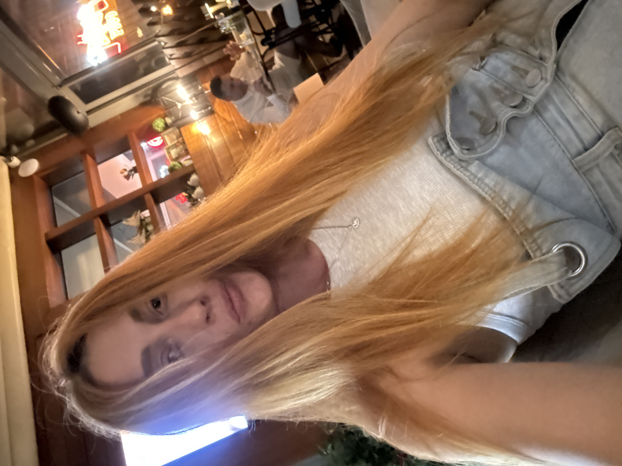 In a dimly lit, wood-paneled establishment, a young woman with long, auburn hair takes a selfie. She is wearing a light-colored tank top and denim overalls. Her hair is partially covering her face, and she is looking towards the camera with a neutral expression. In the background, out of focus, a man in a white shirt and shorts is seated at a table, appearing to be eating. The environment suggests a restaurant or bar with neon signs visible through a window or glass partition. The establishment is located in Budva, Montenegro.