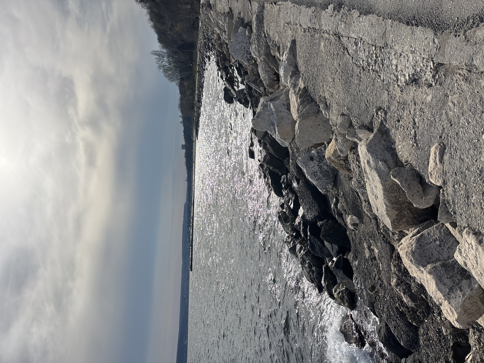 This image captures a view of the Black Sea from the coastline in Varna, Bulgaria. A rocky embankment dominates the right foreground, transitioning from concrete pathway to large, gray and black boulders leading to the water's edge. The sea is shimmering with sunlight, reflecting the sky's gray-white cloud cover. In the distance, a hazy shoreline and some indistinct buildings can be seen along the horizon, suggesting the opposite side of the bay or a distant part of Varna itself. There are no visible people in the frame. The general impression is that of a cold, overcast day by the sea.