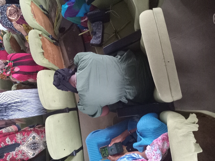 The scene depicts a gathering of individuals seated in rows of light green chairs with black straps, likely in Umueshi, Nigeria. The chairs appear to be upholstered, and some show wear and tear. Several people are seated, with varying attire. One individual wears a green top, black head covering, and a silver necklace. Another person is dressed in blue and pink patterned clothing and is using a cell phone. Other individuals have patterned fabrics or head coverings. The background is undefined.