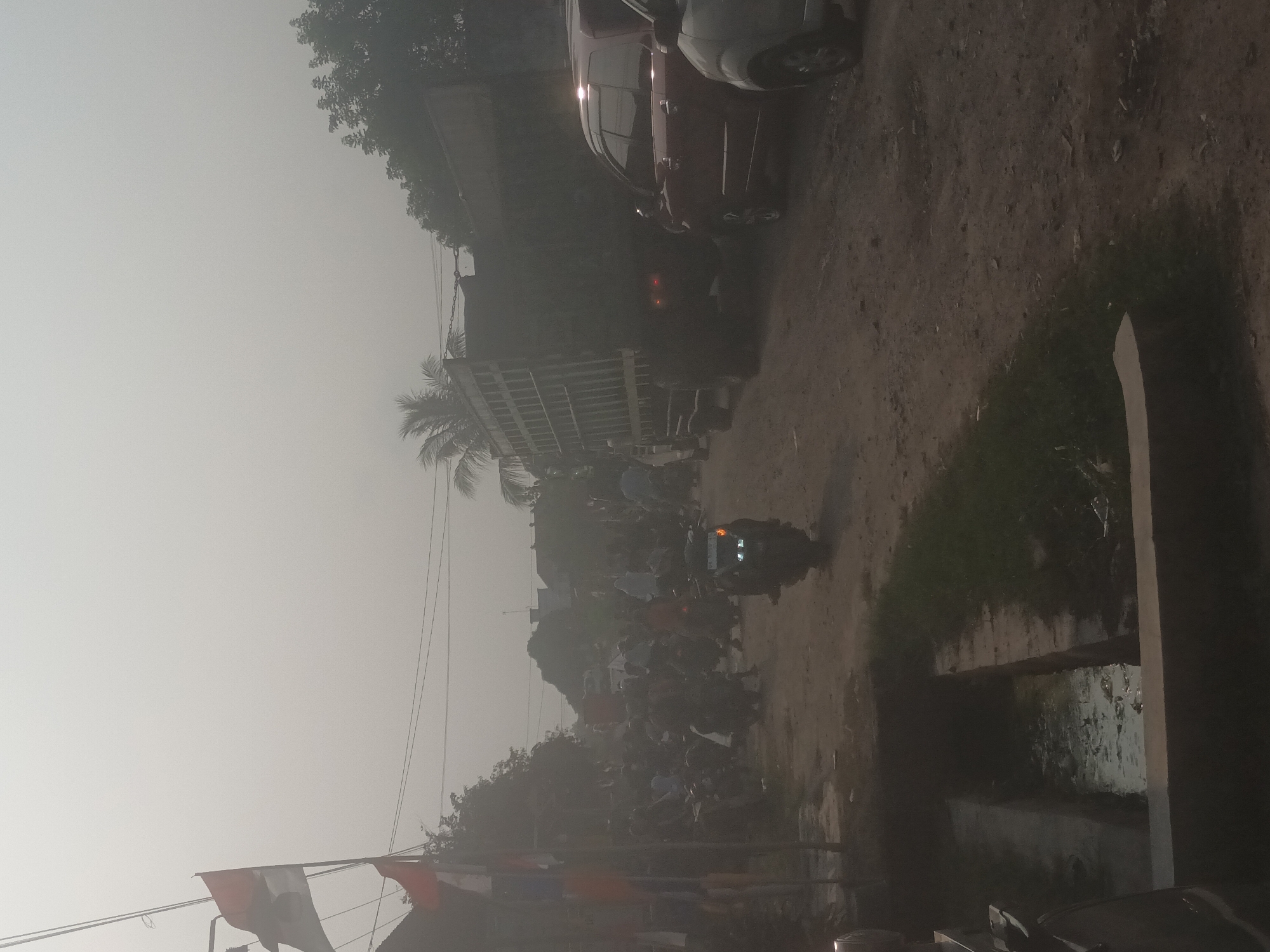The image depicts a street scene in Mulyo Asri, Indonesia, characterized by a hazy atmosphere, possibly due to dust or pollution. A large, dark-colored truck with a caged bed dominates the right side of the frame, parked on a dirt road. To its right, a red SUV is visible. A silver car is partially seen in the foreground on the far right.
On the left side of the road, a group of people are gathered, many appearing to be on motorcycles, suggesting a communal event or a busy intersection. Two flags, one red and white and another with a white emblem on a red background, are prominently displayed on poles in the left foreground, implying a local celebration or gathering. A motorbike with its headlights on is positioned in the middle ground, facing towards the left.
The environment includes a drainage ditch filled with murky water in the lower left corner. Buildings with roofs are visible in the background, interspersed with trees and palm trees. The overall impression is one of a bustling, possibly informal, urban or semi-urban setting.