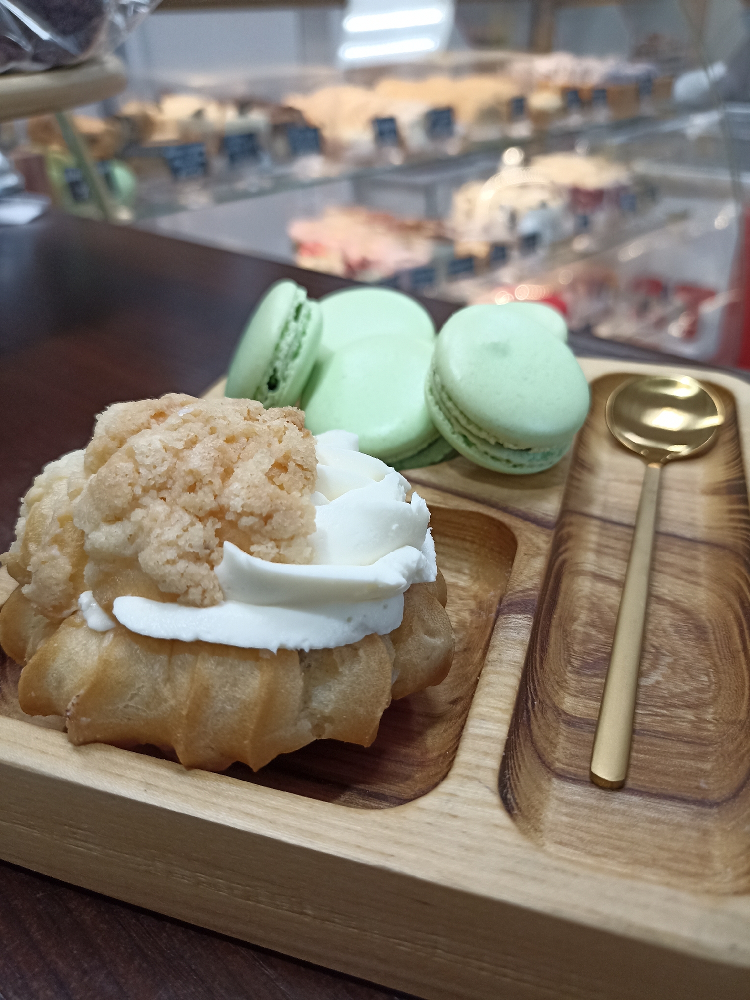 A close-up shot taken in an unknown location in an unknown city depicts a wooden tray containing a cream puff, three green macarons, and a golden spoon. The cream puff is topped with whipped cream and a sugar-crusted topping. The macarons are pale green. Behind the tray, a display case filled with various baked goods is visible. The tray sits on a dark-colored table.
