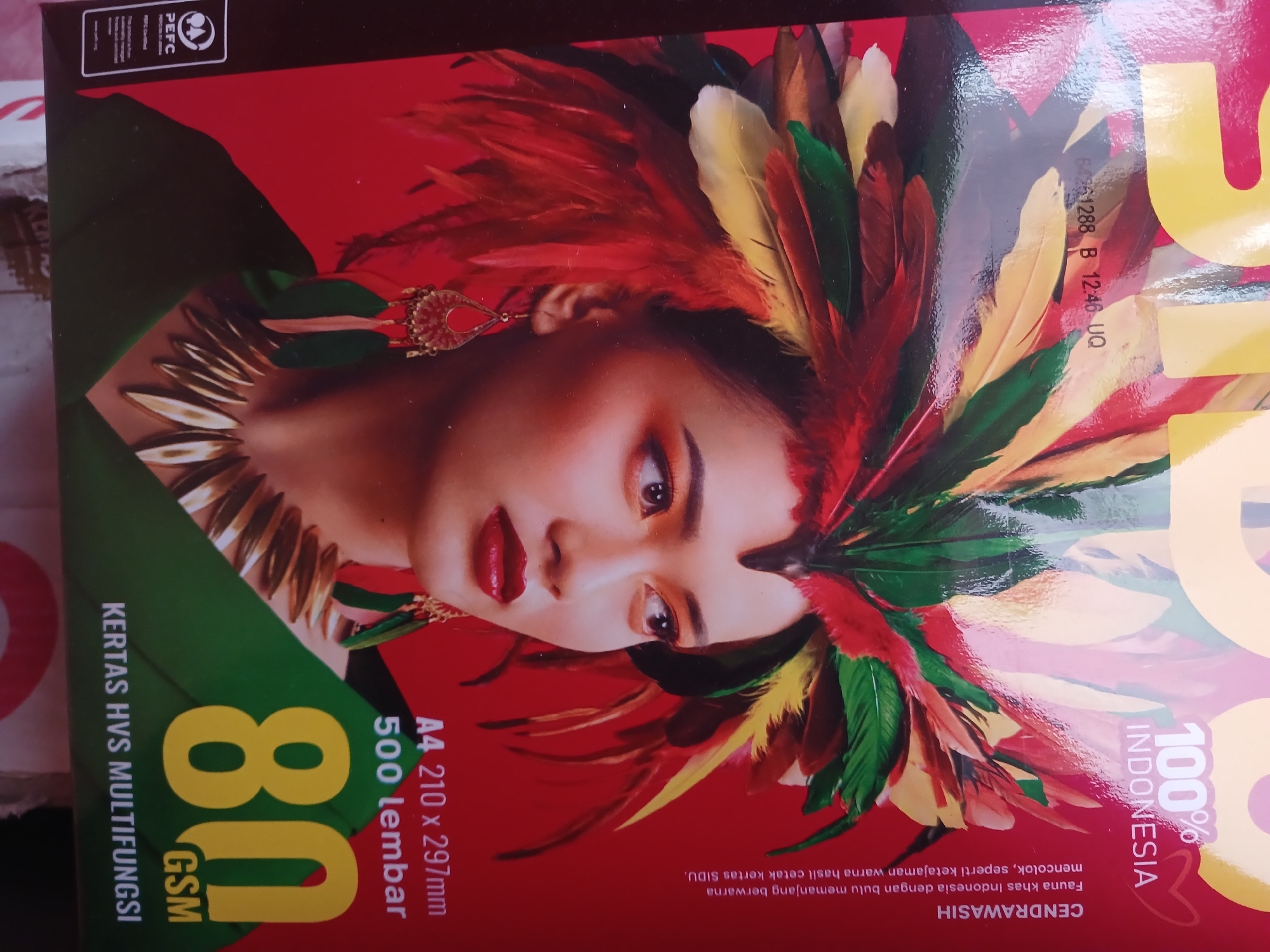 The image shows the front of a red package of paper, possibly for printing. The packaging features a large graphic of a woman's face adorned with colorful feathers, suggesting a vibrant and possibly cultural theme. Text on the package indicates it is "GSM 80" and "500 lembar" (500 sheets), specifying the paper weight and quantity. There are also details about the paper size, "A4 210 x 297mm". The text "KERTAS HVS MULTIFUNGSI" identifies it as multipurpose copy paper. In the lower left corner, text reads "CENTRAWASIH" and describes the "Fauna khas Indonesia" (Indonesian fauna), linking the design to Indonesian natural heritage. The bottom of the package displays "100% INDONESIA". There is no visible information that would allow for the identification of a specific location within Watugede, Indonesia.