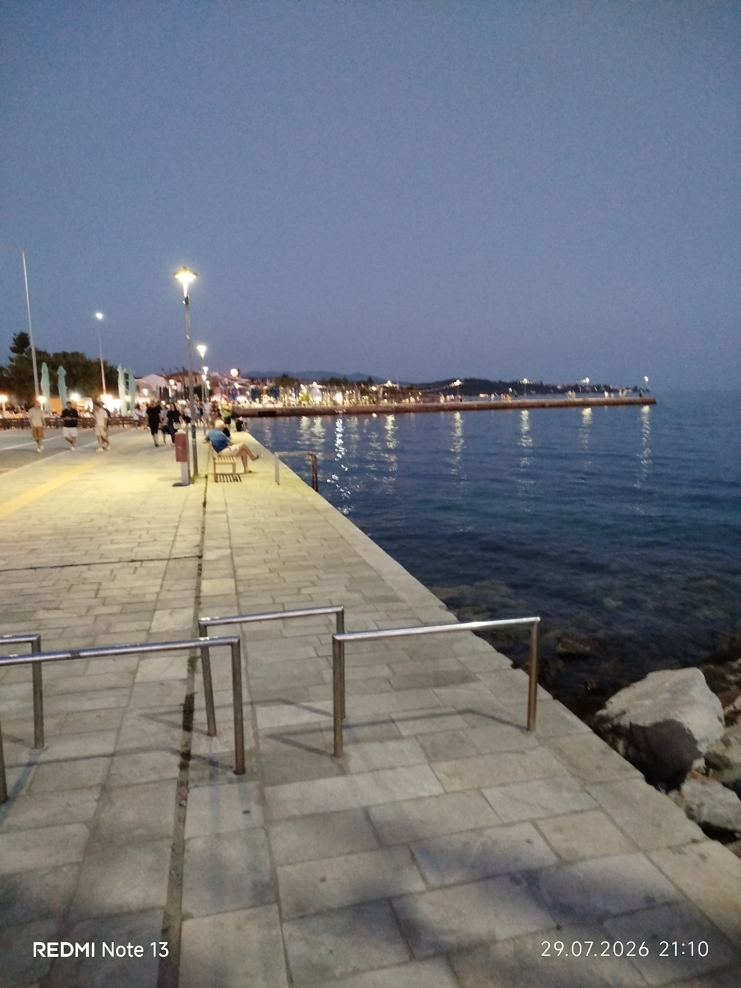 An evening view of a waterfront promenade in Sithonia Municipal Unit, Greece. The promenade is paved with large, grey stone tiles and lined with modern metal railings. The sea is calm and reflects the lights of a distant town that stretches along the coastline. Several streetlights illuminate the promenade. In the foreground, the paved walkway is visible, with shadows cast by the railings. Further down the promenade, a few people are walking, and one individual is seated on a bench, facing the sea. Trees and buildings are visible in the background, along with more lights. The sky is a deep blue, indicating it is dusk or early evening. A rock formation is visible at the edge of the water on the right side of the image.