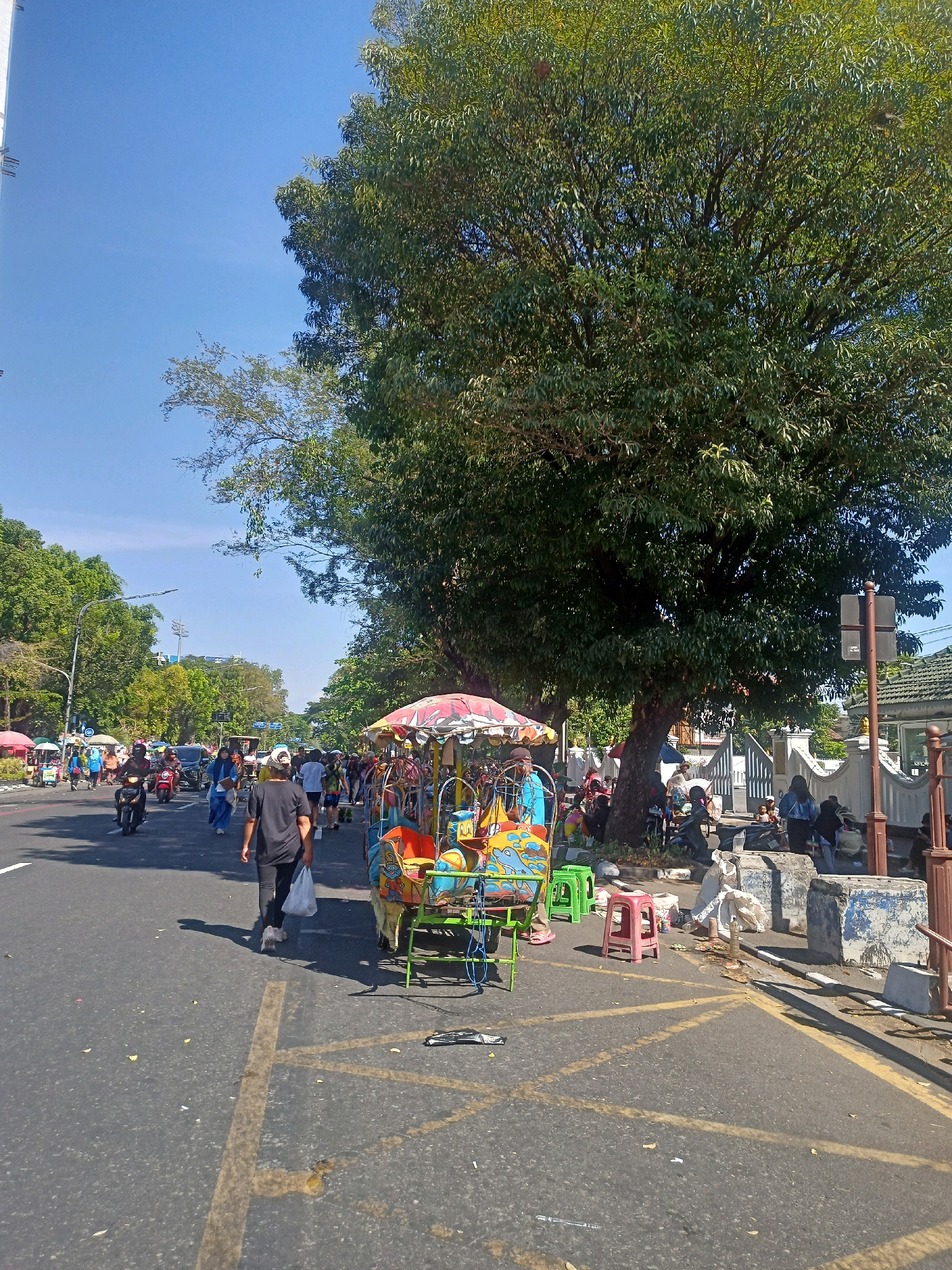 The image depicts a bustling street scene in Surakarta, Indonesia, under a clear blue sky. In the foreground, a colorful pedal rickshaw is parked on the side of a paved road, adorned with a vibrant umbrella and a playful design. Several green and red plastic stools are placed nearby, suggesting a waiting area or a temporary setup for vendors.
The road itself is busy with traffic, featuring motorcycles and cars moving in both directions. Pedestrians are also present, walking along the sidewalk or crossing the street. A man in a black t-shirt and dark pants is walking away from the camera, carrying a white plastic bag.
Lush green trees line the street, providing shade and a natural canopy. On the right side of the image, a white fence and a building with a tiled roof are visible, indicating a more established area. The overall impression is one of a lively urban environment where daily life unfolds amidst a blend of natural and man-made elements.