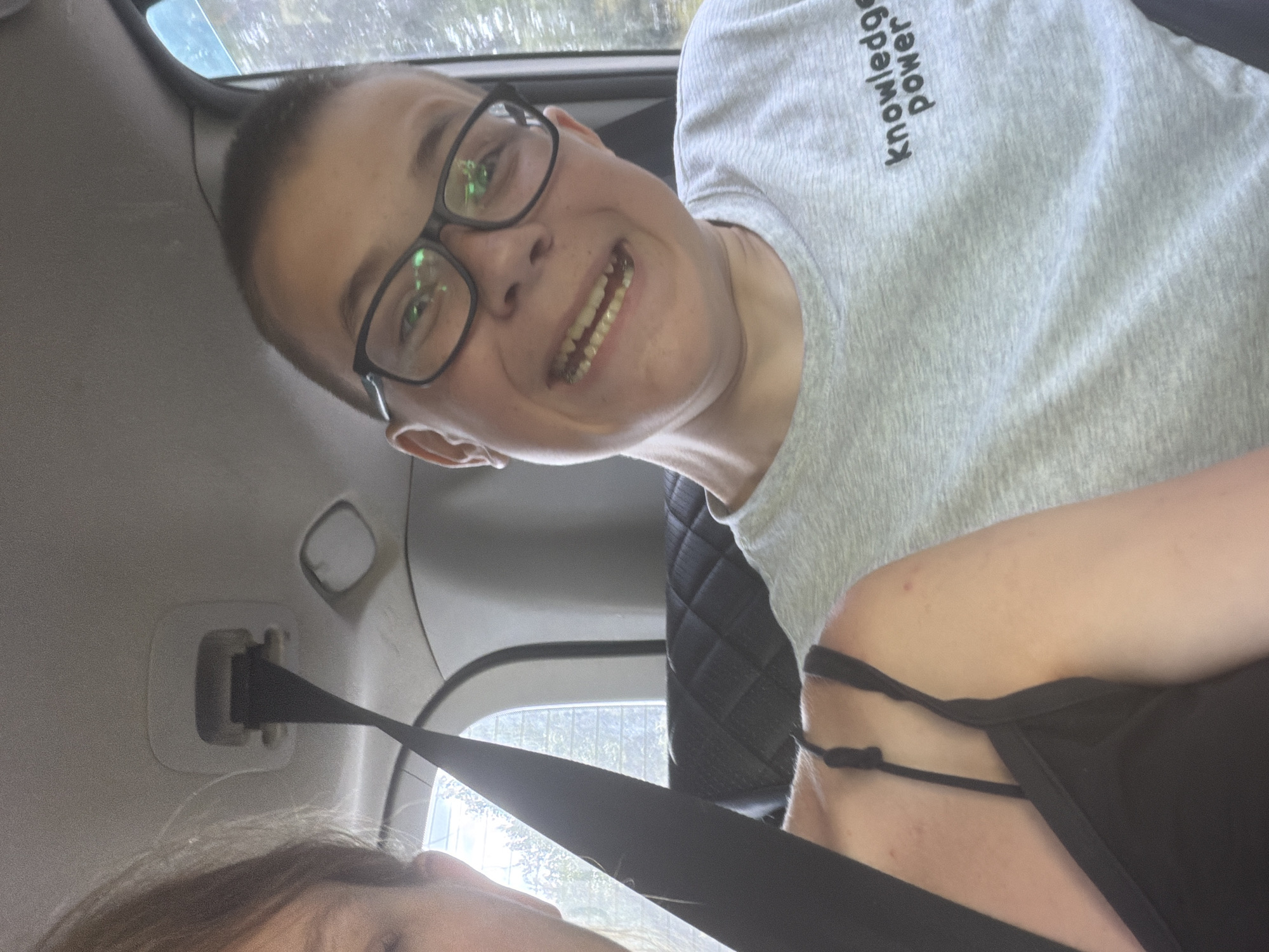 In Novaya Zhizn', Russia, a selfie is taken inside a car. The image shows two individuals, a young person with glasses and braces, smiling widely, and another person, whose face is partially visible on the left side of the frame, with their shoulder and arm also in view. Both individuals are wearing seatbelts, indicating they are traveling in the vehicle. The interior of the car is visible, including the ceiling, a black upholstered seat, and a window through which trees and foliage can be seen. The young person is wearing a grey t-shirt with the text "knowledge power" printed on it. The scene appears to be a casual moment captured during a car journey.
