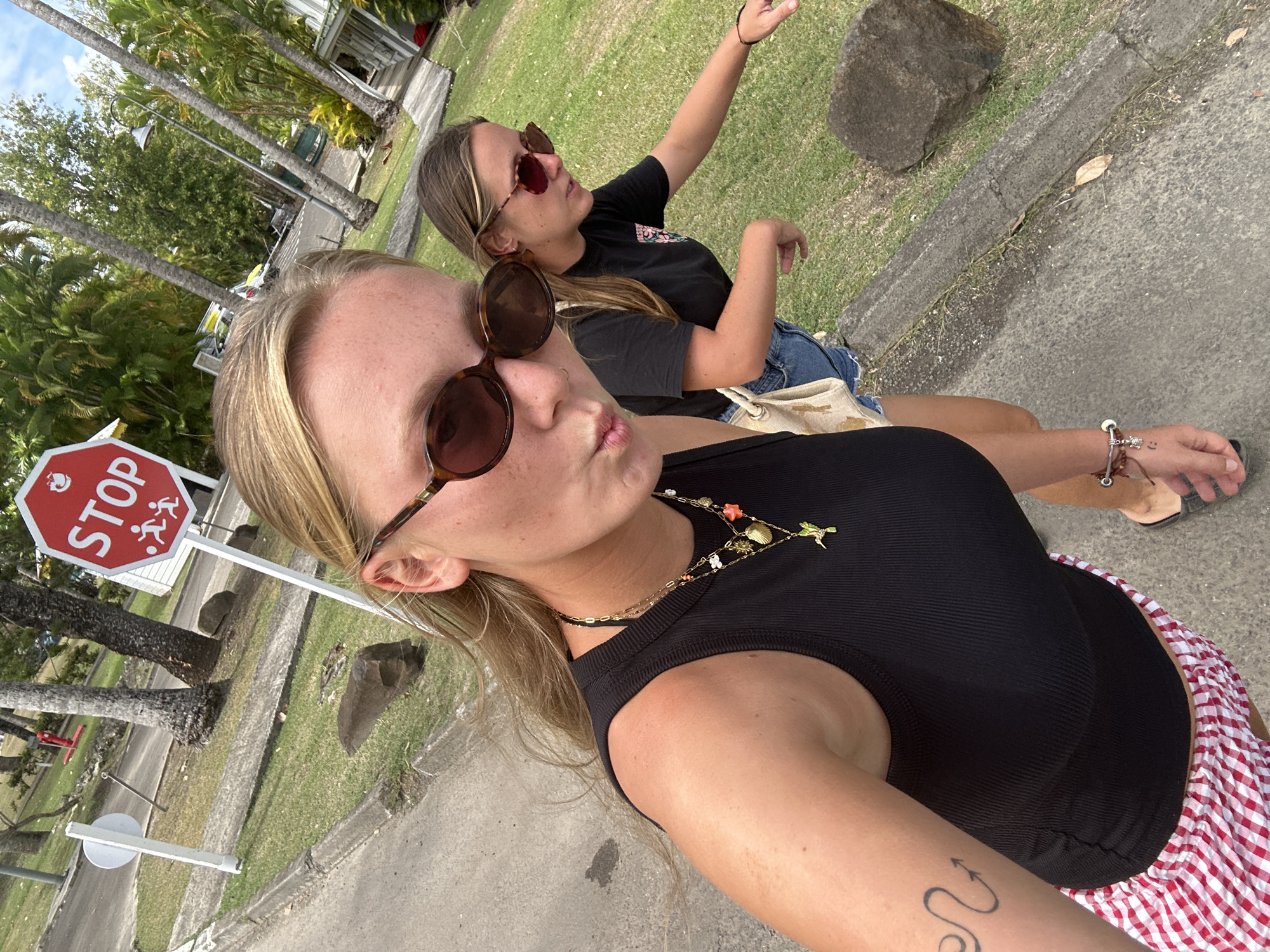 Two young women are posing for a selfie on a sidewalk in Sainte-Anne, France. The woman in the foreground, with blond hair, is wearing a black tank top, red and white checkered shorts, and sunglasses. She has a necklace and a tattoo on her left arm. The woman in the background, with brown hair, is wearing a black t-shirt, denim shorts, and sunglasses. She is sitting on a ledge with her right arm raised. A stop sign is partially visible to the left of the foreground woman. A grassy area with trees and a building are visible in the background.
