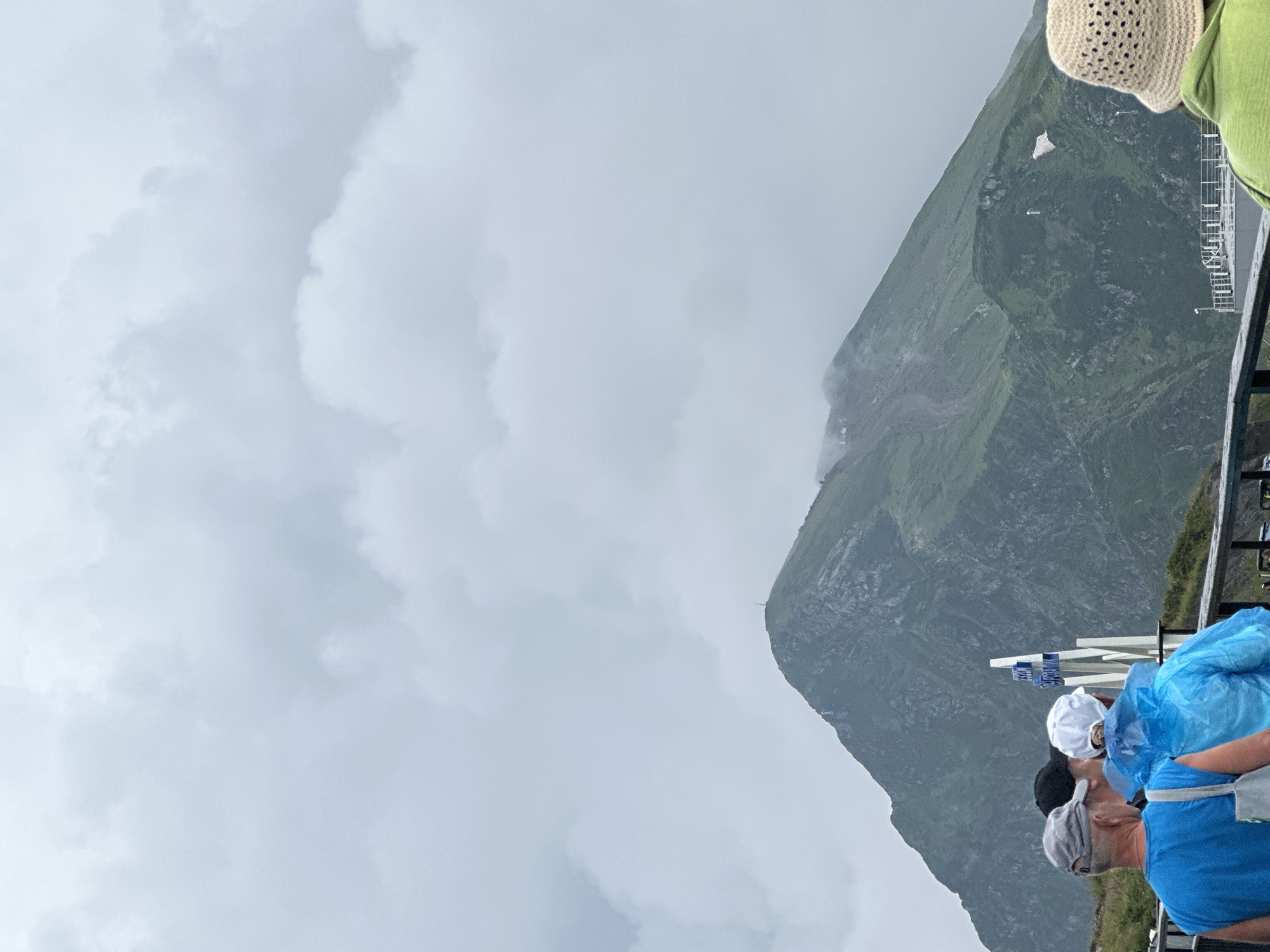 The image shows a view of a mountain in Estosadok, Russia, under a cloudy sky. In the foreground, three people are visible from the back, standing on a viewing platform with a railing. The person on the left wears a blue t-shirt and a grey cap. The person in the middle is wearing a black cap and the person on the right is wearing a blue raincoat and a white cap. A white signpost with blue lettering is positioned between the group and the mountain. The mountain itself is covered in lush green vegetation, with some rocky outcrops visible. A path or track can be seen winding up the side of the mountain. The summit of the mountain has a small structure, possibly a radio tower or observation post. The overall atmosphere of the image is overcast and slightly hazy, suggesting a cooler, possibly damp, day in the mountains.