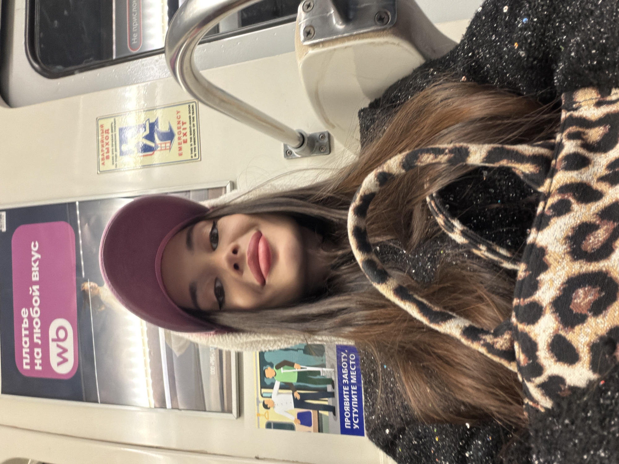 The image is a selfie taken by a woman on a public transport vehicle, likely a metro train. She is wearing a pink hat and a black jacket with a leopard print bag.
The interior of the train car is visible in the background. There are advertisements and informational signs in Russian, including an "EMERGENCY EXIT" sign. A sign shows images of people and a message to "show care and give way," probably referring to offering seats to those in need. Another sign advertises "clothes for every taste". A Wildberries (wb) logo appears on the advertisement.
Based on the Russian signage, the image was likely taken in Russia. Given the interior design, architecture and signage it is Saint Petersburg.