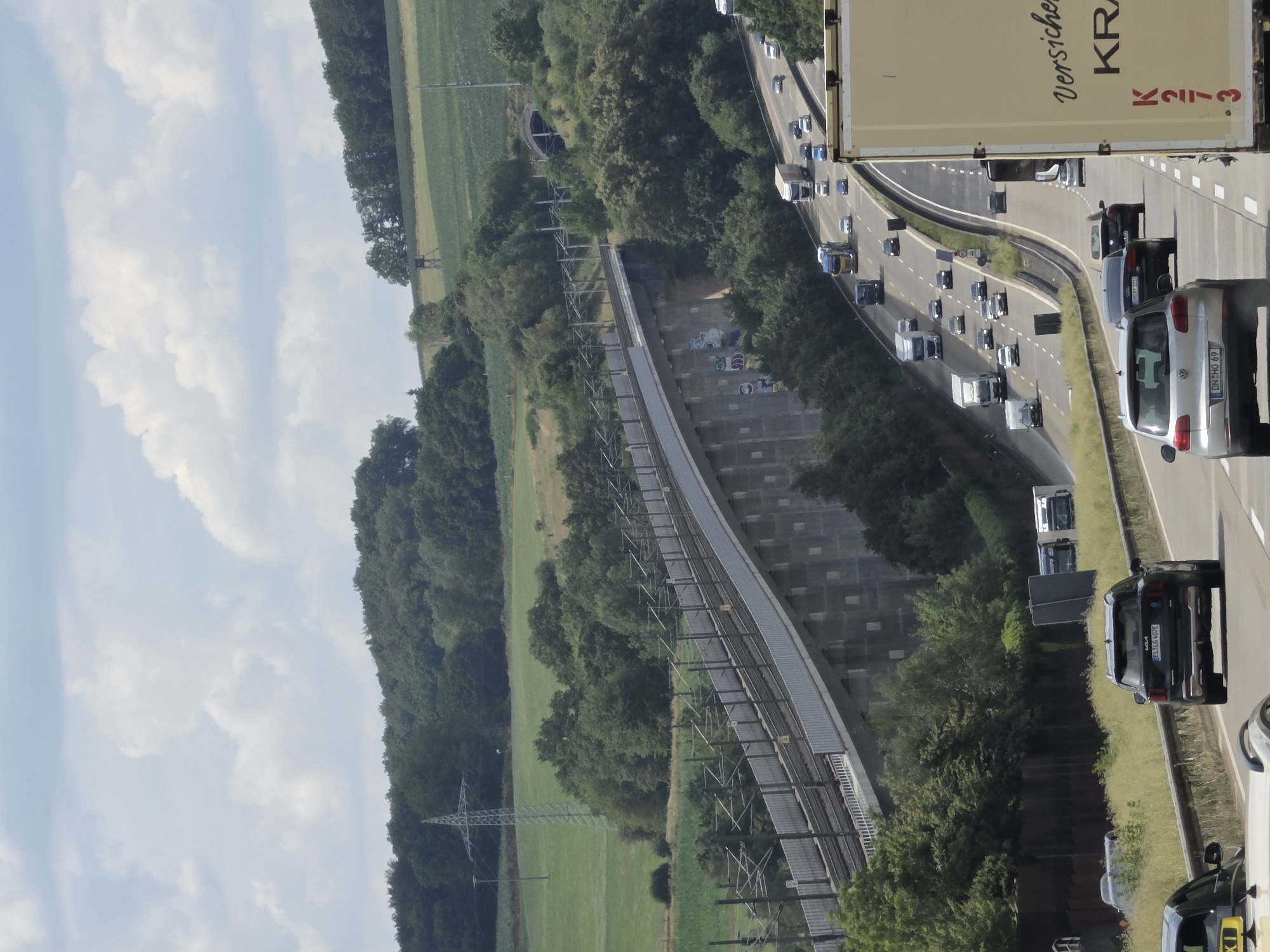 This image depicts a wide, multi-lane highway in the rural landscape near Neustadt (Wied), Germany. The highway curves through rolling hills covered with dense green trees and fields. In the foreground, several vehicles, including cars and trucks, are traveling on the highway. A large semi-trailer truck with "Versicher..." and "KRA" visible on its side is prominently positioned in the right foreground. Further up the highway, a dark SUV with a license plate ending in "407E" is visible. In the middle ground, a significant elevated railway bridge with overhead power lines traverses the landscape, supported by concrete pillars. A tunnel entrance is visible in the background amidst the trees. The sky above is partially cloudy with a mix of blue sky and white clouds. The overall scene conveys a sense of transportation infrastructure integrated with a natural environment.