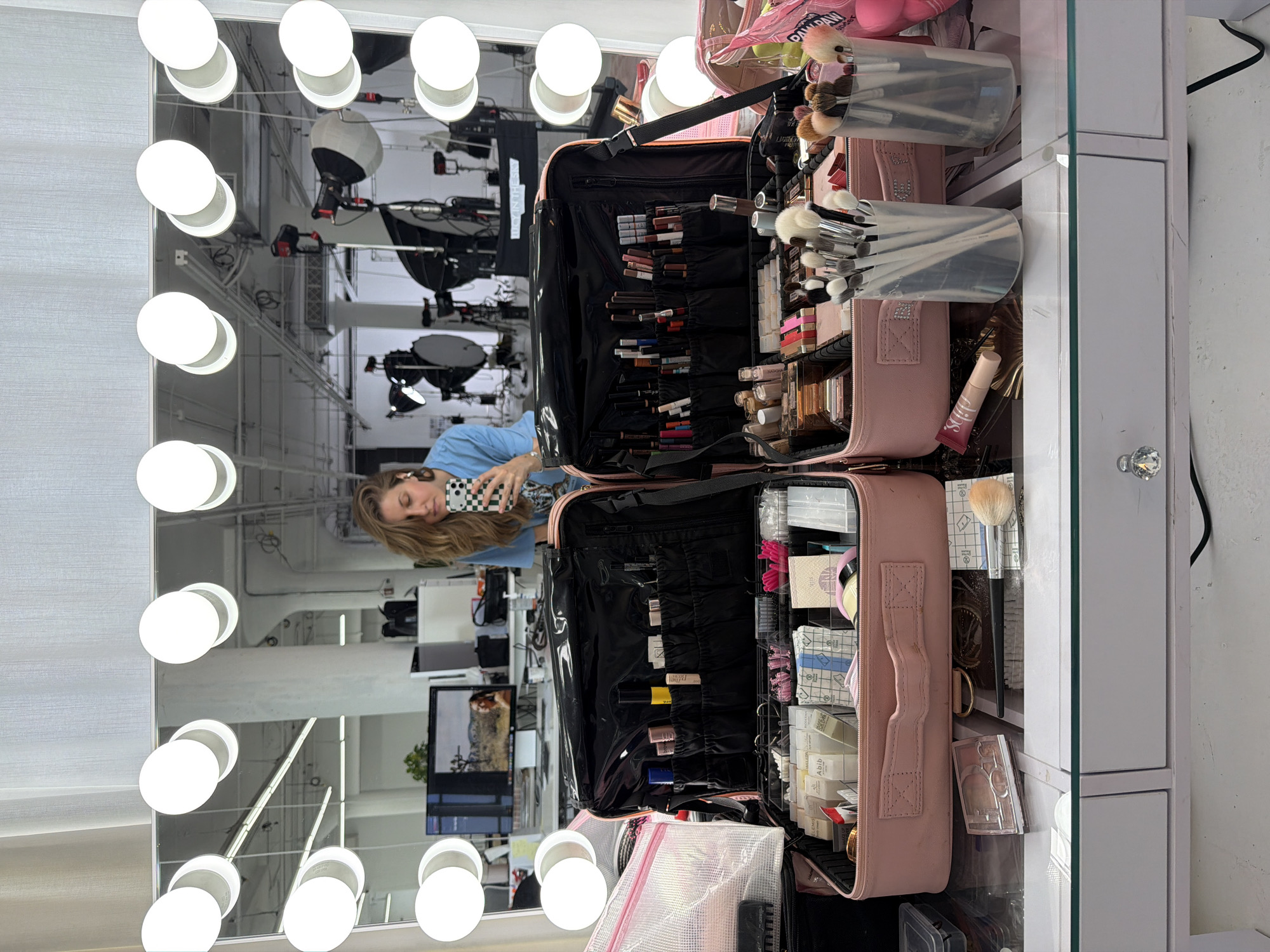 A reflective, rotated image shows a woman taking a selfie in a lighted makeup mirror. The woman has light brown hair and is wearing a blue top, holding a phone with a checkered case. The reflection reveals a studio-like environment, likely in Los Angeles, USA, based on the typical studio setups and equipment visible. Overhead, there are numerous studio lights and lighting rigs. The foreground is dominated by a pink makeup case filled with various cosmetic items, brushes, and tools. The vanity table is white with a decorative knob on the drawer. The mirror is surrounded by many illuminated light bulbs.
