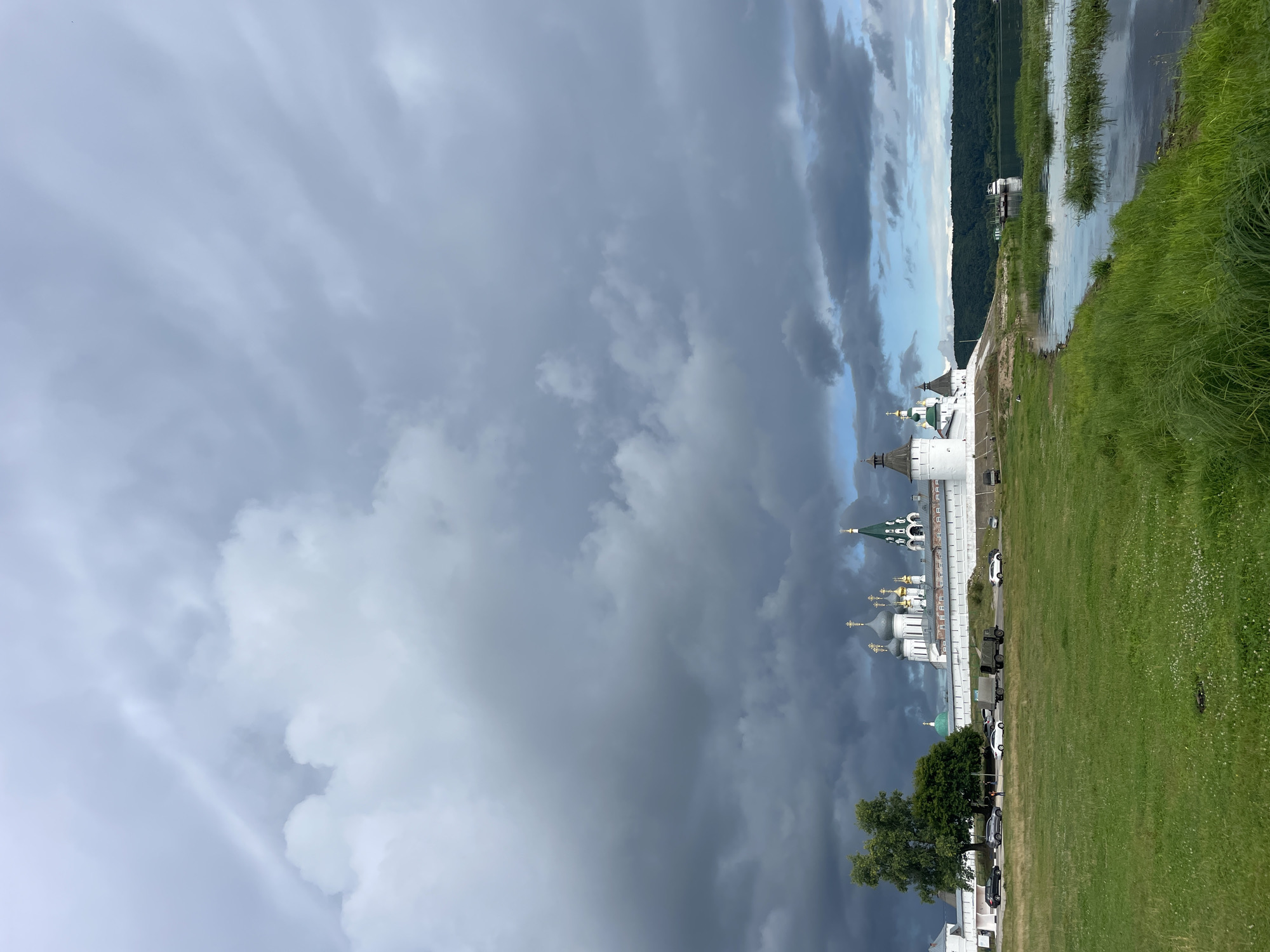 The image shows a view of Makar'evo, Russia. In the foreground, a grassy field with some scattered white flowers slopes down to the edge of a calm body of water. The water reflects the sky and the vegetation along its banks. In the middle ground, a white monastery with numerous domes and towers stands on a slightly elevated embankment. The monastery has white walls and distinctive green and gold domes, some topped with crosses. Several cars and a military truck are parked along a road in front of the monastery. A large, lush green tree stands to the left of the monastery. In the background, a densely forested hill rises on the opposite side of the water. The sky above is filled with dramatic, dark gray storm clouds, with patches of lighter sky visible in the distance. A few boats are docked near the shore on the far side of the water. The scene depicts a historical and religious site under a brooding sky.