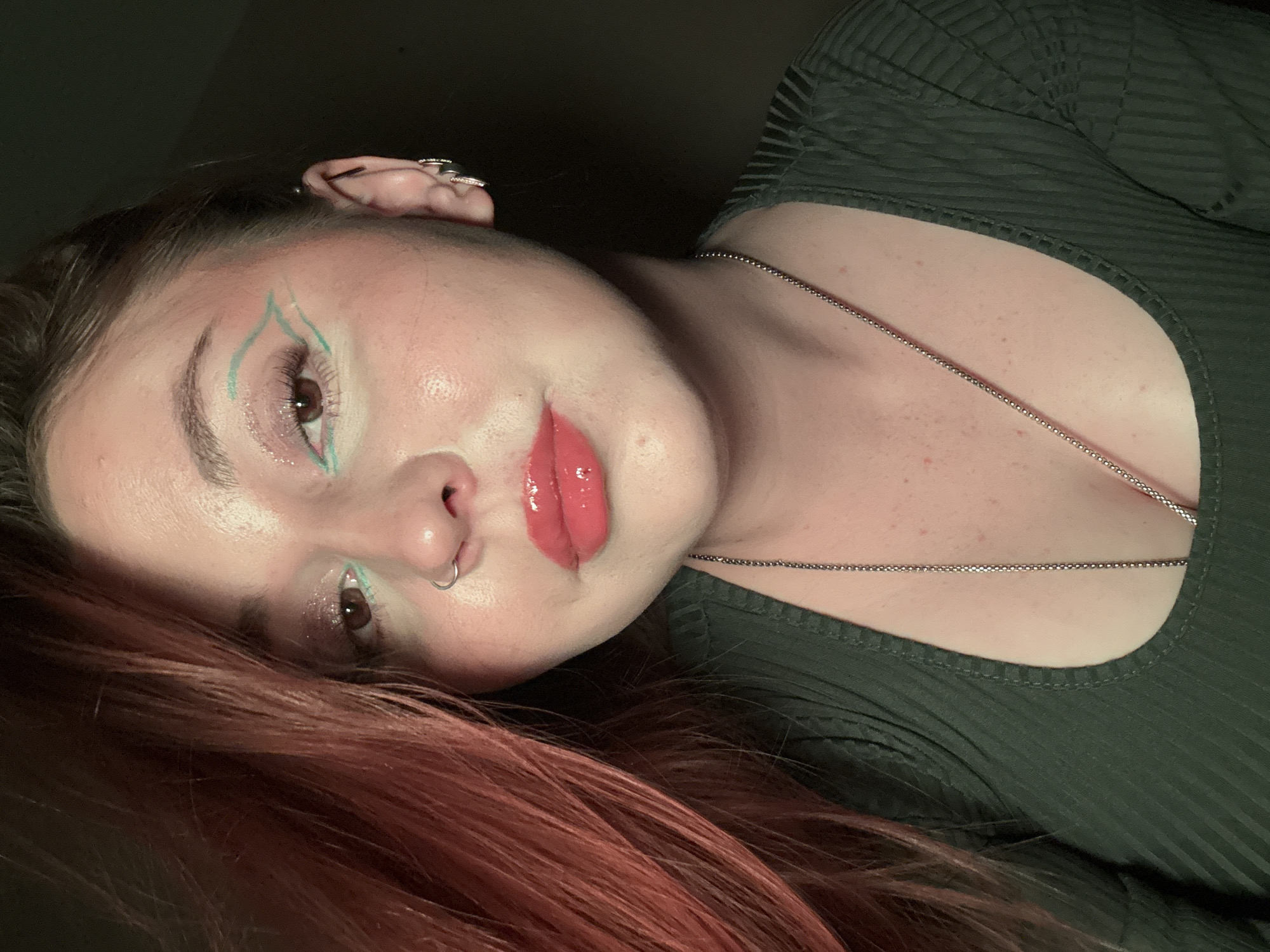The image is a close-up portrait of a woman with reddish-brown hair. She is wearing a dark green ribbed top and a silver chain necklace. She has a nose ring in her left nostril and jewelry in her left ear. Her makeup includes iridescent eyeshadow, turquoise eyeliner accentuating her eyes and brows, and glossy red lipstick. The background is dark, and there is no evidence to suggest the photo was taken in Izhevsk, Russia.