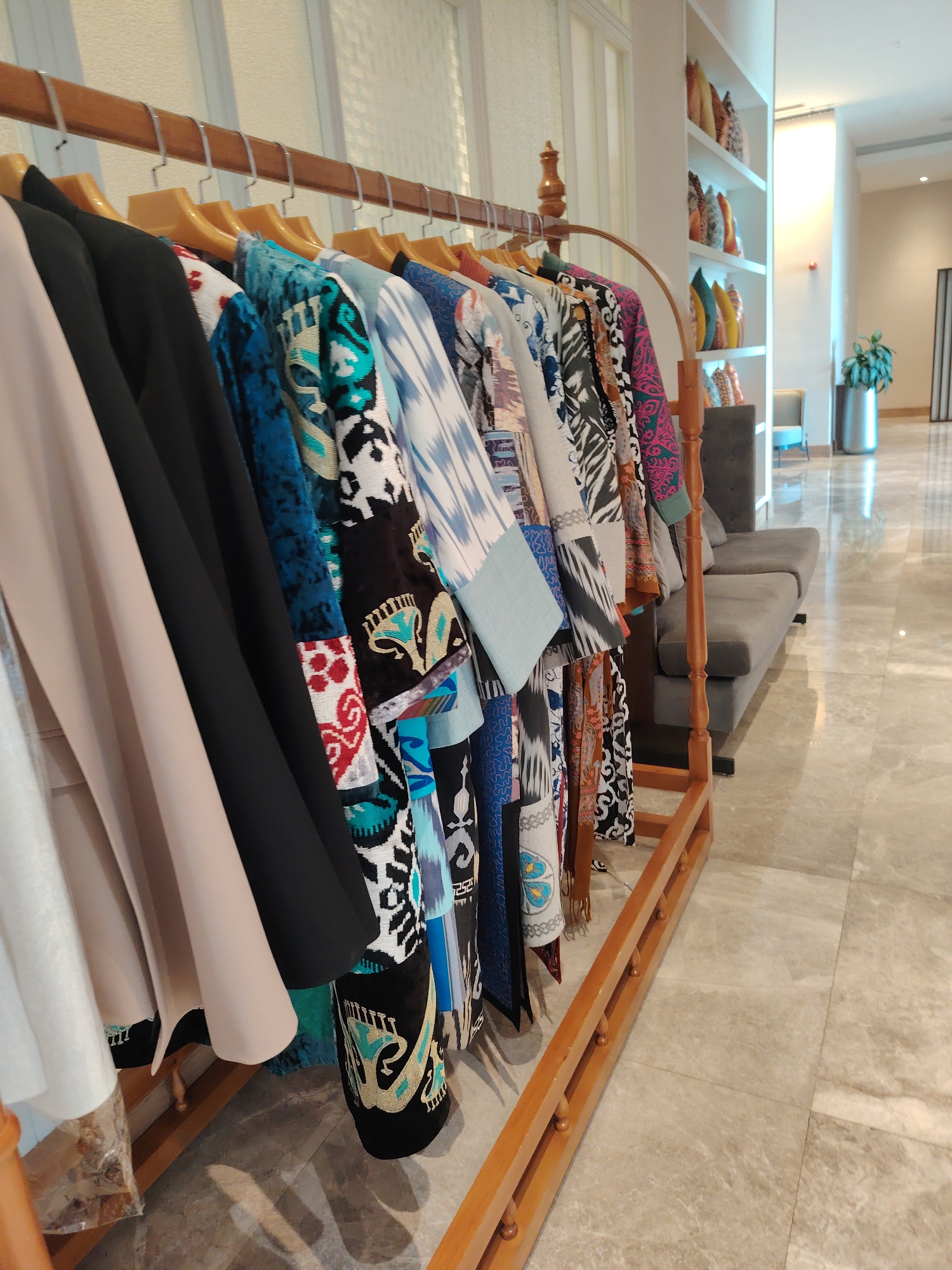 This image depicts a retail display of colorful, patterned garments hanging on a wooden rack in an indoor setting. The garments feature intricate designs with vibrant colors such as blue, teal, pink, and white, suggesting a cultural or artisanal origin. Several of the garments appear to be made of rich fabrics, possibly silk or velvet, with embroidery and appliqué work.
The background shows a well-lit interior with tiled flooring and a sofa. Shelving units filled with decorative pillows are visible in the background, further suggesting a retail or showroom environment. The overall ambiance is clean and organized.
The location, Kuchtumgali, Uzbekistan, is known for its rich textile traditions and vibrant handicrafts, which aligns with the visual evidence of the garments displayed. The scene captures a segment of what could be a boutique or a stall within a larger market or gallery, showcasing the region's unique fashion and textile art.