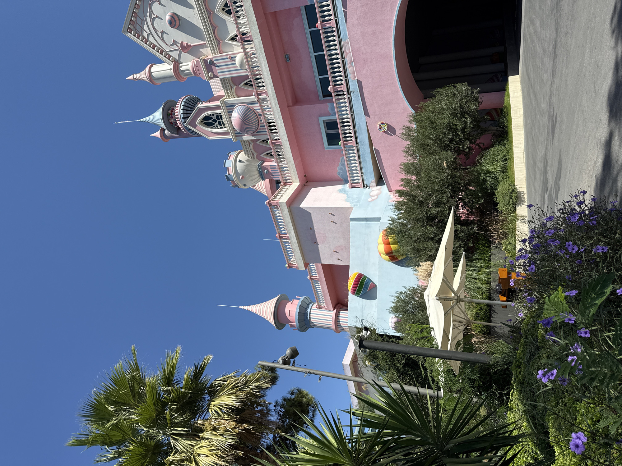 The image shows a brightly colored, whimsical castle-like building in Bodrum, Turkey. The structure is primarily pink with blue and white accents, featuring multiple towers, spires, and decorative elements such as balconies and arched doorways. Several hot air balloon decorations are visible on the exterior walls. In the foreground, lush green foliage and vibrant purple flowers create a natural setting. Two large white umbrellas are set up on what appears to be an outdoor seating area. The sky is clear and blue. The overall scene suggests a theme park, resort, or similar entertainment venue designed with a fairytale aesthetic.