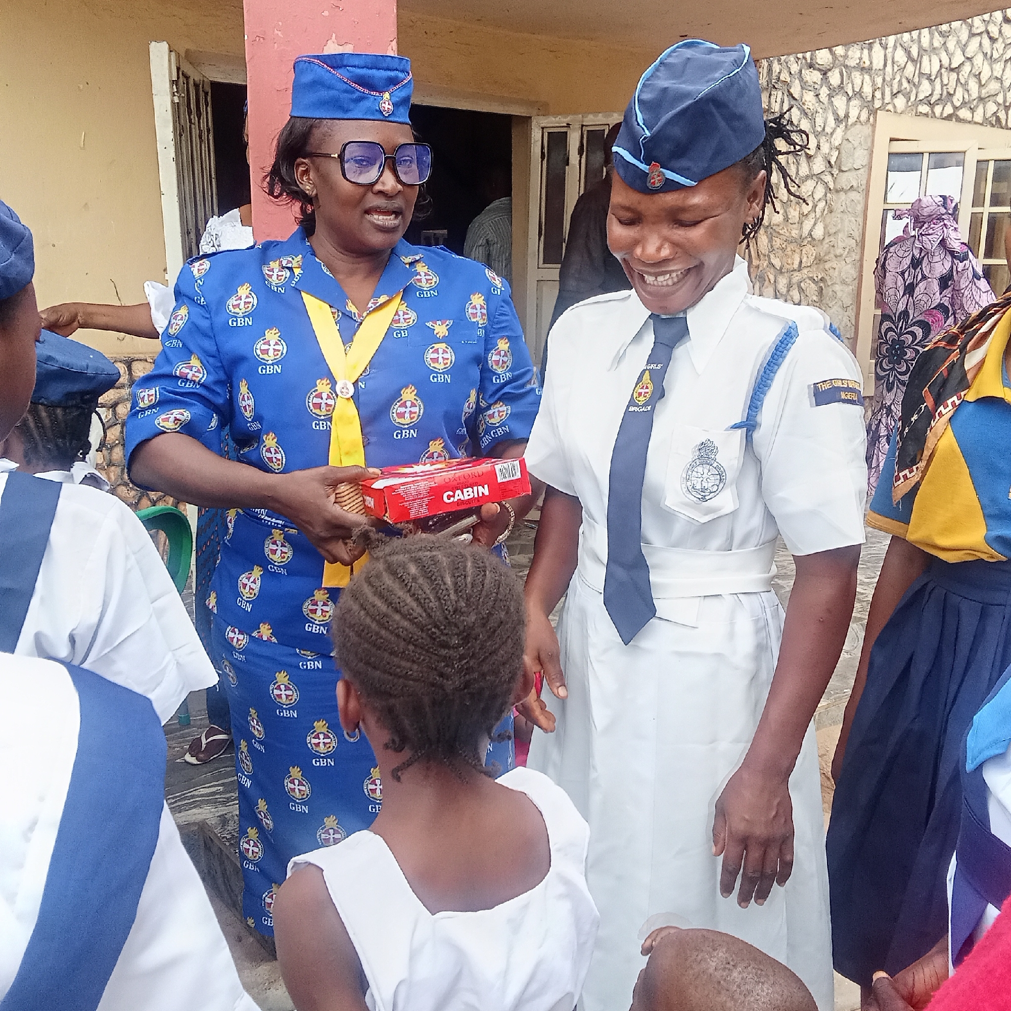The image shows two women in uniforms, likely associated with a youth organization, surrounded by several children. The woman on the left wears a blue patterned shirt with the acronym "GBN" repeated throughout, a matching blue hat, and glasses. She is holding a red box of "CABIN" biscuits. The woman on the right is smiling, wearing a white uniform with a navy blue tie and hat, and a badge that says "GIRLS BRIGADE". The children are of varying heights and wear white or blue and yellow clothing, suggesting they are part of the same or a related group. The background shows a building with a mix of stucco and stone or brick siding. The setting appears to be outdoors, possibly in a city or urban area given the presence of buildings. The city and region of Unknown, Unknown is not present.