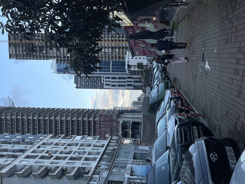 A street lined with parked cars is seen between modern buildings, some under construction. Two people are walking along the sidewalk, passing a wall with graffiti. The sky is visible between the structures. Trees hang from the upper left corner. Car license plates are visible in the foreground.