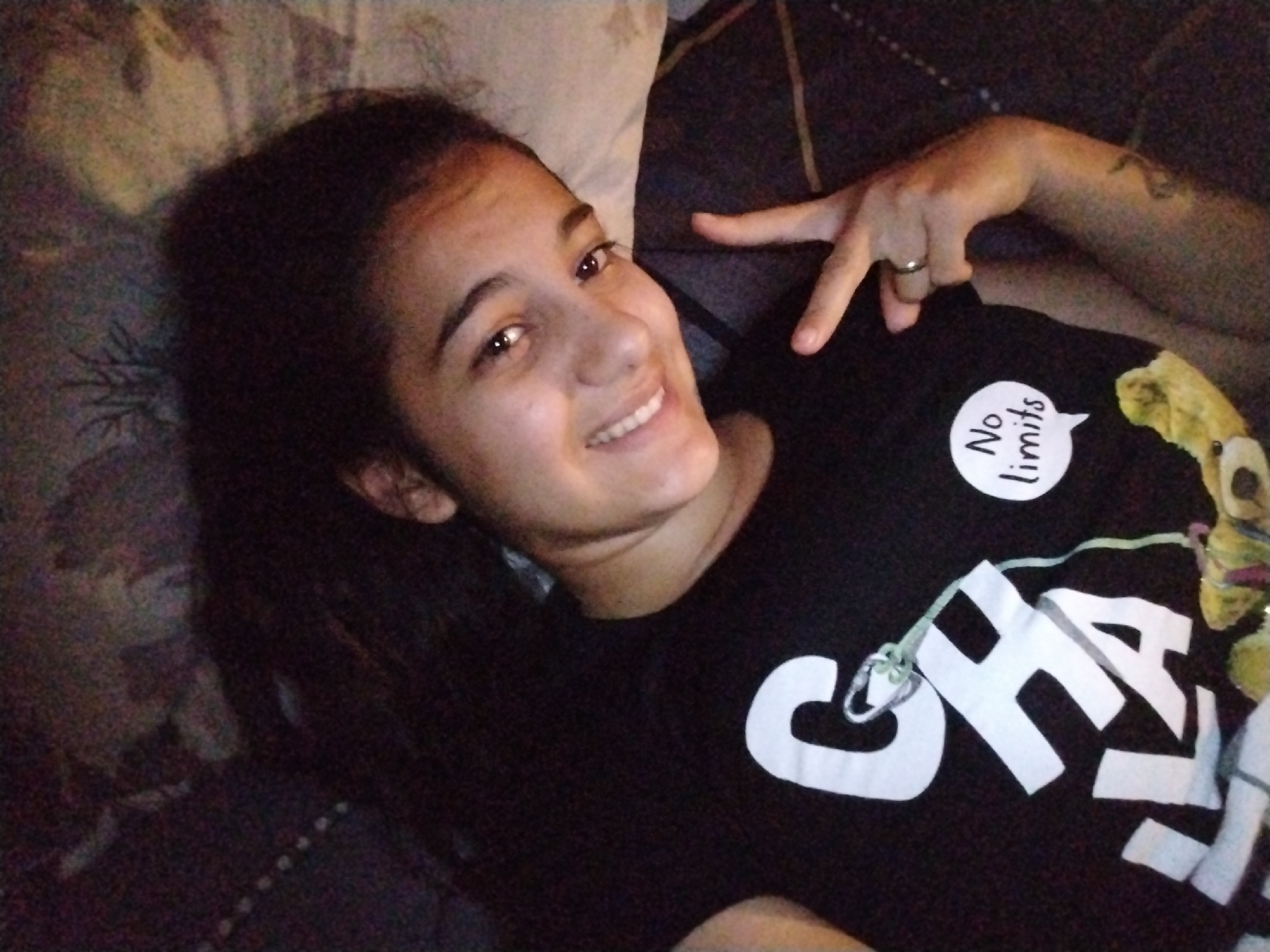 A young woman with dark hair is lying down, smiling directly at the camera. She appears to be in an indoor setting, possibly on a bed or sofa, indicated by a patterned pillow or cushion behind her head and to her left. Her head is resting on the surface, and she is looking upwards towards the camera.
She is wearing a dark-colored t-shirt. Her right arm is bent, and her hand is visible, making a "V" sign or peace sign with her index and middle fingers extended. A simple, silver-colored ring is worn on the ring finger of her right hand. A faint tattoo is visible on her right forearm/bicep area.
The lighting is subdued, giving the image a slightly grainy appearance, suggesting it might be evening or taken in low light. The woman's expression is joyful and relaxed.
On her t-shirt, there is a white speech bubble with the text "No limits" written inside it in black lowercase letters. Below this, large white uppercase letters are visible, spelling "CHA" and possibly more letters of a word that is cut off or obscured. A thin green string or lace detail is tied around part of the design, near the letter 'C'.