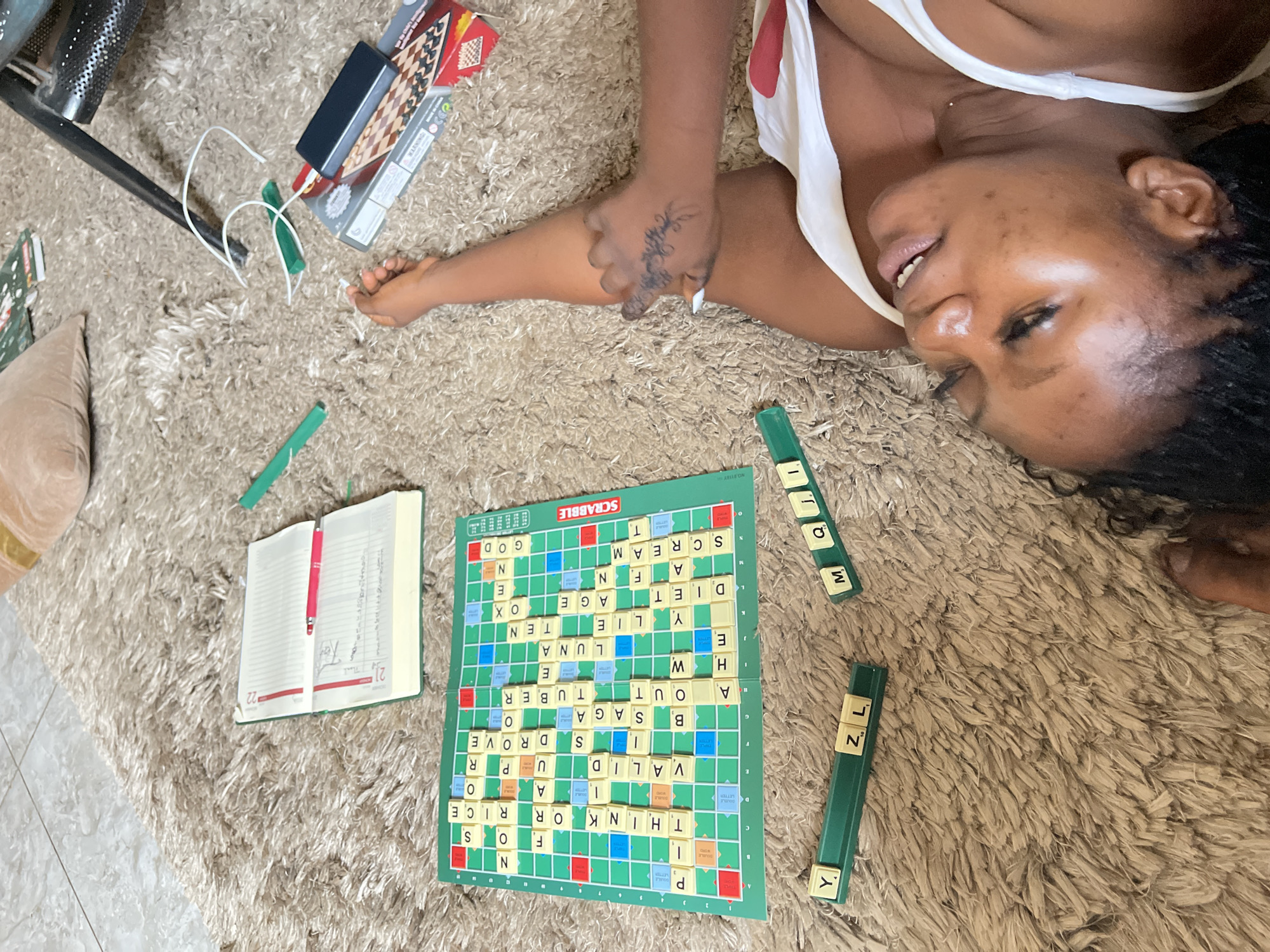 A person is seen from an overhead perspective, lying relaxed on a shaggy beige rug, seemingly playing or having just played Scrabble in an indoor setting in Kuchingoro, Nigeria. The individual, a woman with dark skin and braided hair, is on her back with her head turned to the left, smiling serenely. She wears a white tank top, and a tattoo is visible on her left hand.
The primary activity revolves around a game of Scrabble, with a green Scrabble board laid out on the rug. Numerous letter tiles spell out words like "GOD," "OX," "AGE," "TEN," "LUNA," "TUBER," "SAGA," "DROVE," "VALID," "THINK," "RICE," "SCREAM," "DIET," "LIE," and "OUT." Two green tile racks are also present, holding various letters including 'Y', 'L', 'Z', 'M', 'Q', 'J', 'I'. An open notebook with a red pen rests nearby, displaying numbers "22" and "21" on its pages, possibly for scorekeeping or notes.
In the background, a beige throw pillow, a black metallic chair leg, and a stack of board game boxes (one appearing to be a chess/checkers set) are visible, along with white charging cables. The overall scene suggests a leisurely and comfortable atmosphere, highlighted by the woman's relaxed posture and pleasant expression. Visible text includes "SCRABBLE" on the board, "DOUBLE LETTER" on game squares, and the numbers "22" and "21" in the notebook.