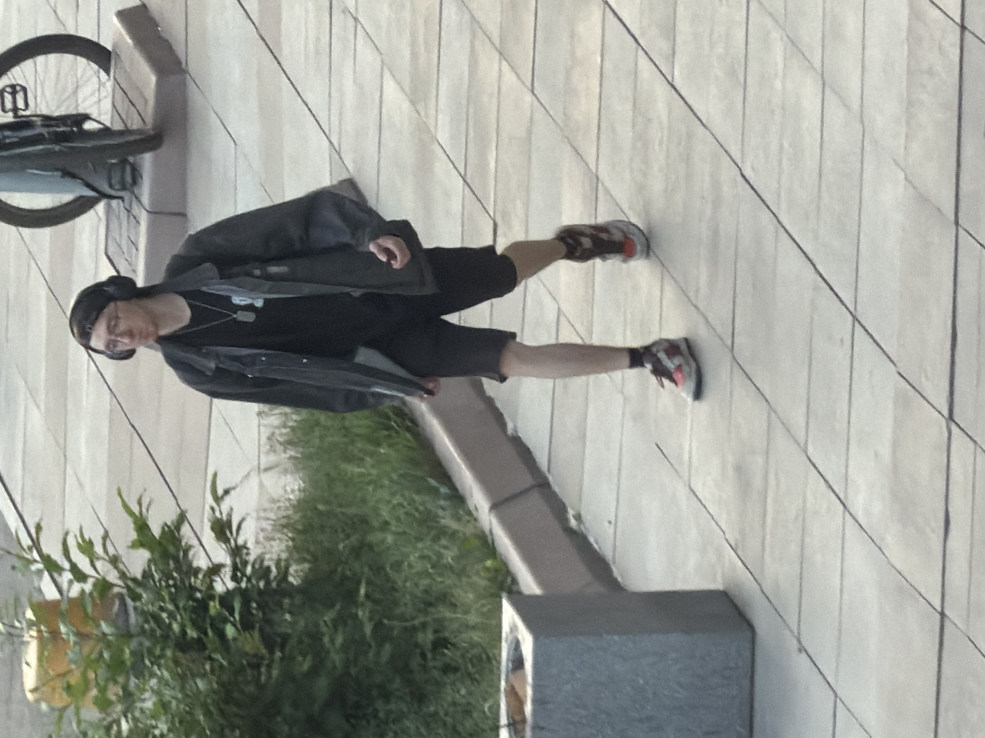 A person walks on a paved sidewalk, wearing headphones, a black t-shirt, black shorts, and a dark jacket. The person is also carrying a backpack and wearing athletic shoes. There is a planter to the left of the frame with green foliage. In the background, a bicycle is parked next to a stone base. The scene appears to be an urban environment, possibly in Krasnoyarsk, Russia, given the request.