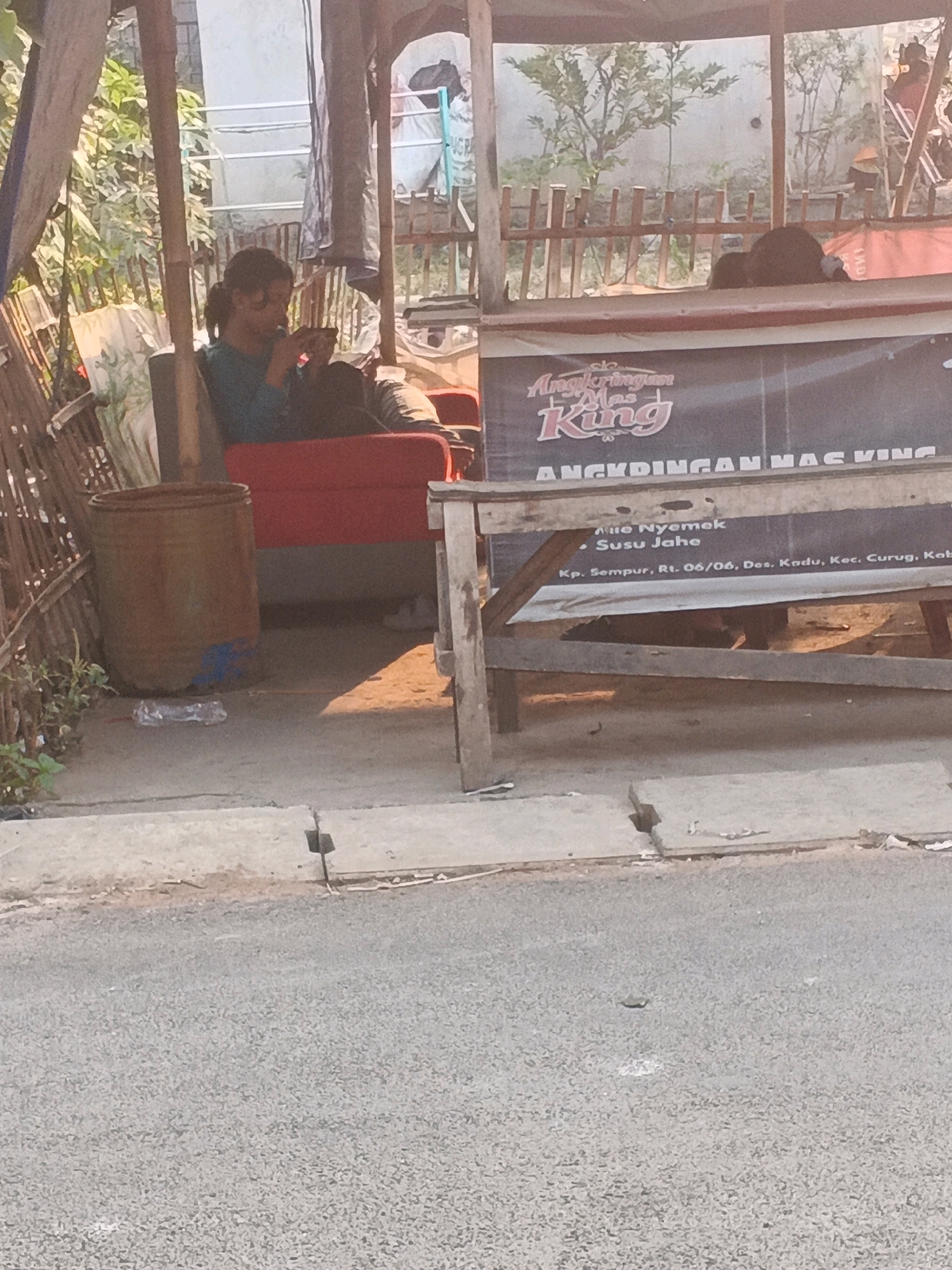 The image depicts an outdoor scene in Kadu, Indonesia, likely a roadside food stall or small shop. In the foreground, a paved road transitions to a concrete walkway. Behind the walkway, a makeshift stall is visible, constructed with bamboo poles and wooden planks. A large banner with Indonesian text and a logo featuring the word "KING" is prominently displayed on the stall.
A person, appearing to be a young woman with dark hair pulled back, is seated on a red sofa inside the stall, looking down at a mobile phone. Her attire is a blue-green shirt. Next to her, a large, cylindrical metal container, possibly a trash can, stands on the ground. A plastic bottle lies on the ground near the container.
The background shows vegetation, a wooden fence, and parts of buildings. The lighting suggests it is daytime, with sunlight casting shadows. The overall impression is a humble, functional outdoor establishment.