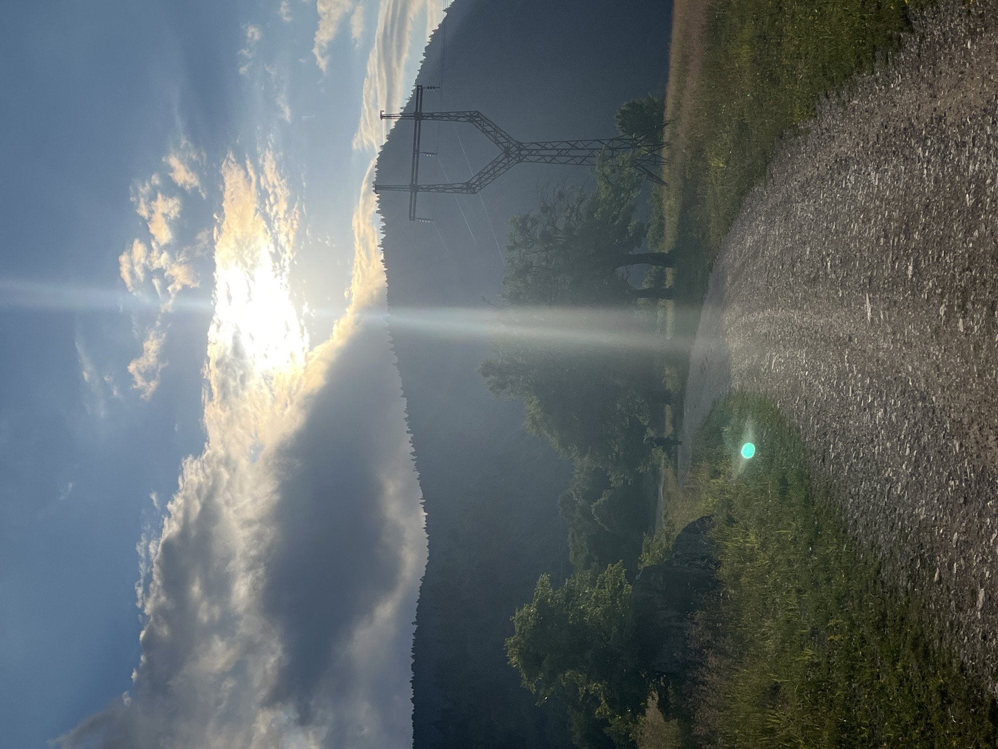 The image depicts a wide, unpaved dirt road curving through a grassy meadow with scattered trees. In the background, a heavily forested mountain slopes upwards, its peak obscured by heavy clouds. A tall, metal power line tower stands to the right of the road, silhouetted against the sky. The sun is partially visible through the clouds, casting a bright lens flare down the center of the image. The overall scene is one of a rural landscape, possibly in the vicinity of Nikortsminda, Georgia, characterized by natural elements and a single man-made structure. There are no discernible interactions or actions taking place.