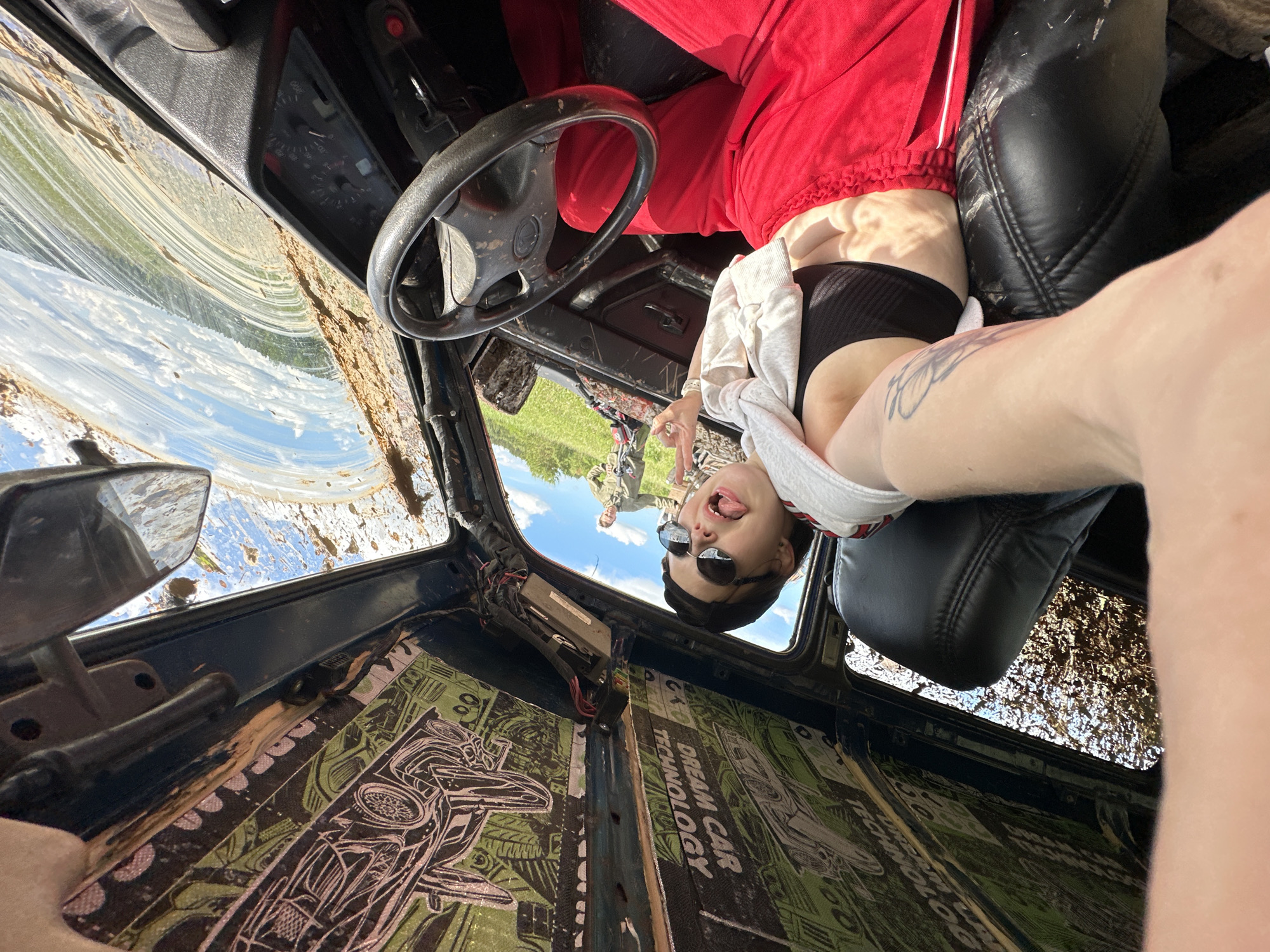 The image shows the interior of a blue vehicle, possibly a car, with extensive damage and mud splattered on the windshield. The interior paneling has custom designs featuring car illustrations and the phrase "DREAM CAR TECHNOLOGY." A person wearing red pants and a black top is in the passenger seat, with a white garment draped over her. The person is leaning back in the seat and making a peace sign with their right hand. The steering wheel is black and worn. In the background, visible through the open roof, are two additional people standing outside on a grassy area, possibly in Ilyinsky, Russia.