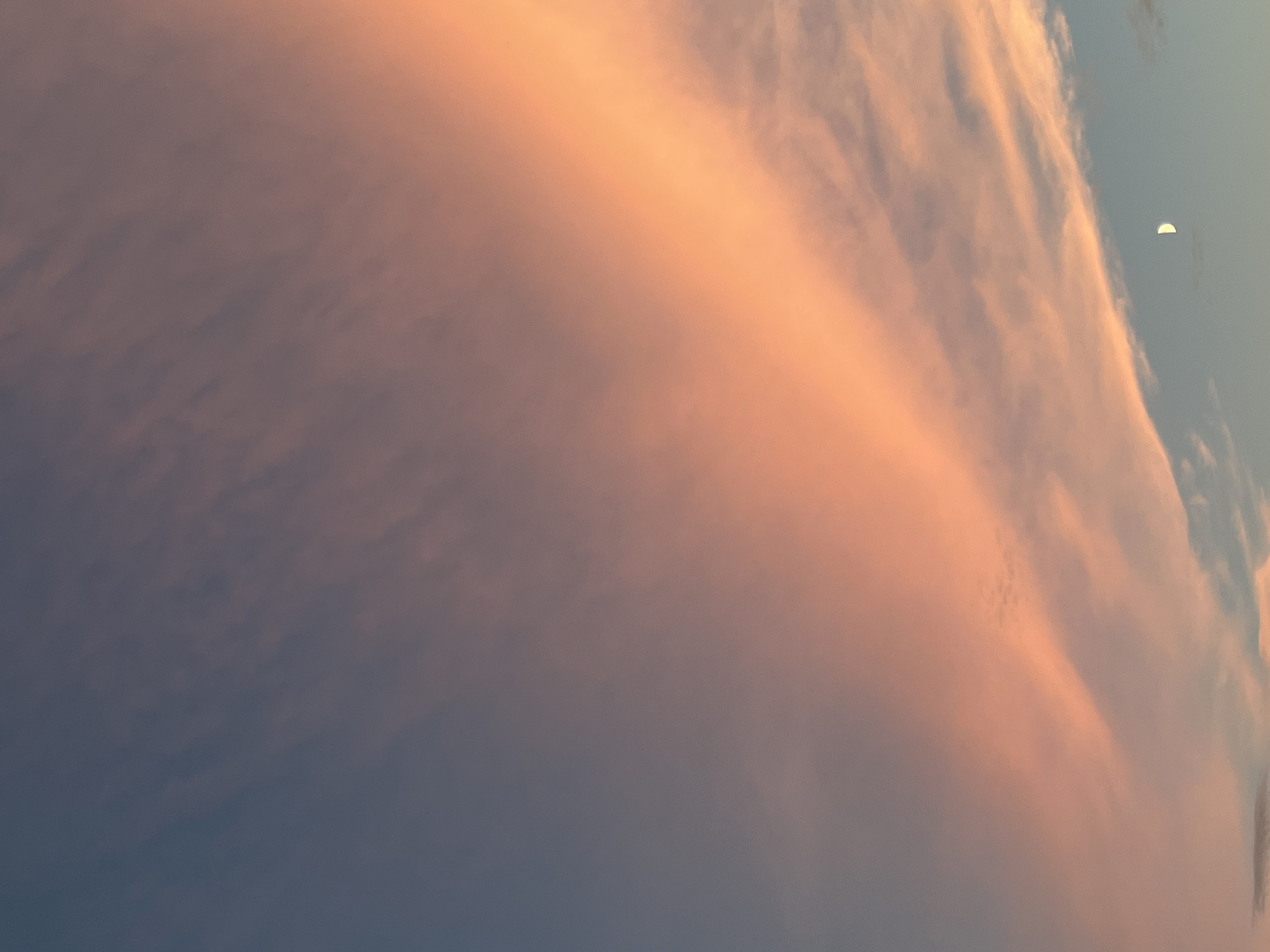 The image depicts a twilight sky over Saint Petersburg, Russia. A sliver of the moon is visible in the lower right quadrant, partially obscured by wispy, pink-tinged clouds that stretch diagonally across the frame. The sky transitions from a deep blue at the top to lighter shades of blue and pink towards the horizon. There are no people or man-made structures visible in the image. The scene is serene and atmospheric, characteristic of a sunset or sunrise in a city with clear skies.