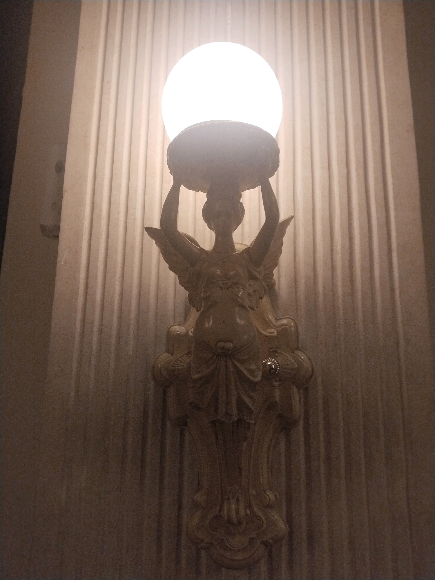 A close-up, frontal, low-angle shot captures a vintage-style wall sconce featuring a sculpted angelic figure holding a frosted glass globe light fixture. The figure, made of a light-colored material, has wings and is depicted from the waist up, wearing draped clothing. The globe emits a bright white light, illuminating the ribbed wall behind it. The wall appears to be made of a textured material with vertical grooves. The overall lighting is dim, with the sconce being the primary light source. The image does not contain any people or other identifiable objects or activities. There is no information in the image to suggest its location in Sinduadi, Indonesia.